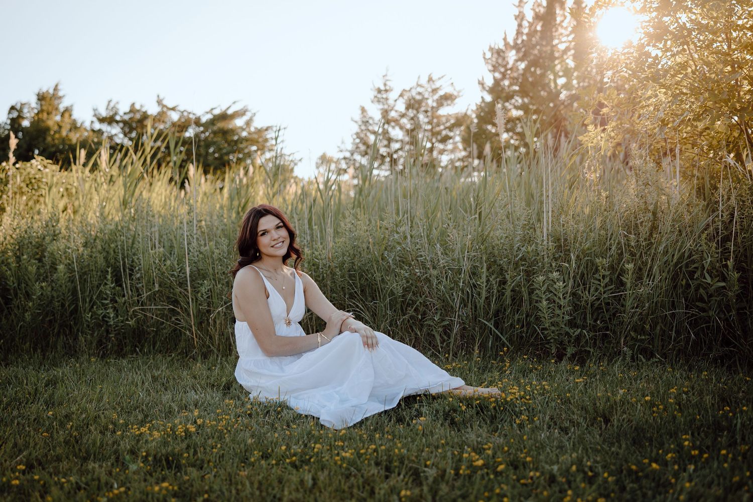 Western Nebraska Senior Session girl in a white dress sits gracefully on green grass with tall reeds and golden sunlight behind her.