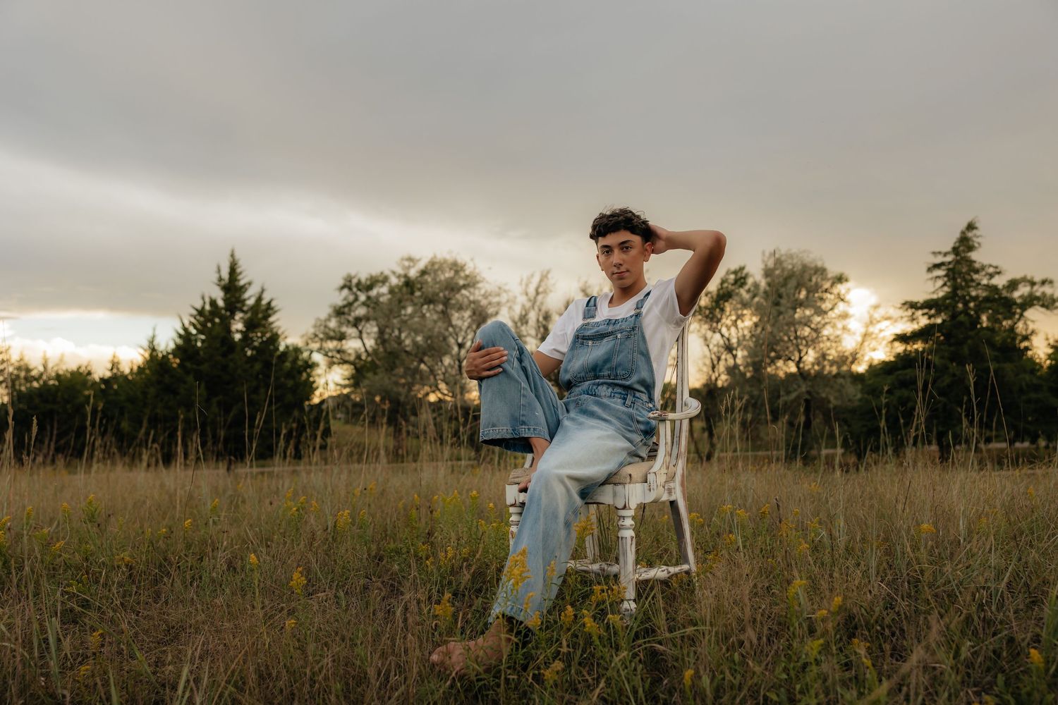 North Platte Nebraska Senior Session young man sitting on white chair in golden field at sunset, wearing denim overalls and white shirt.