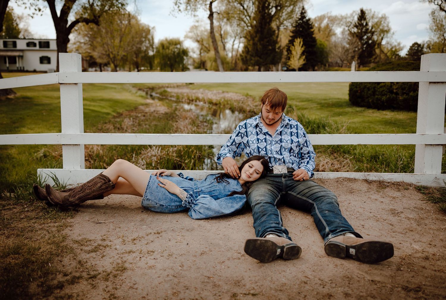 A Western Nebraska Couple Session couple sits along a white fence with woman's head laying on mans lap