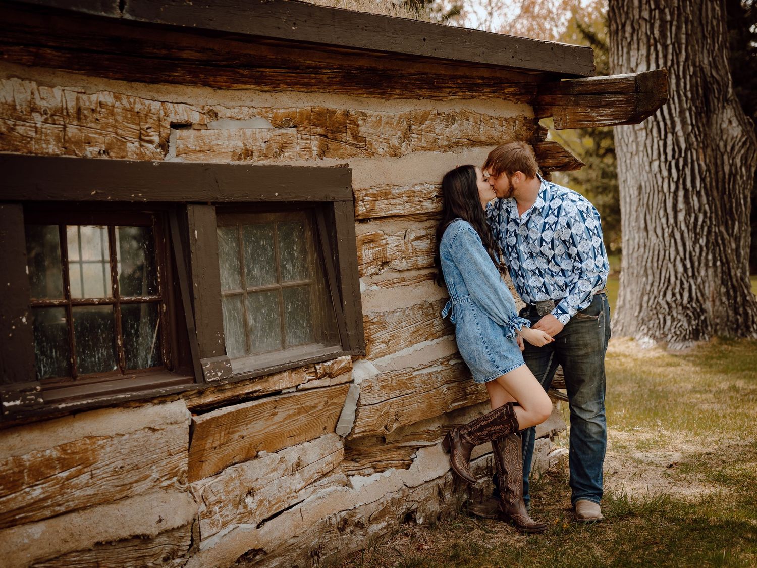 Western Nebraska Couples Session Couple sharing a romantic kiss leaning against a rustic sod house wall outdoors near a large tree.