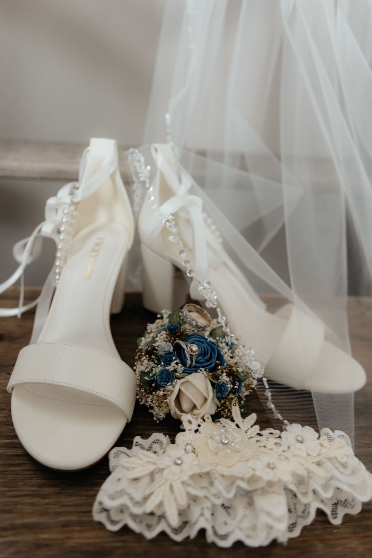Western Nebraska Wedding heels with pearl straps, lace garter, and blue floral boutonniere arranged on wood with a wedding veil backdrop.