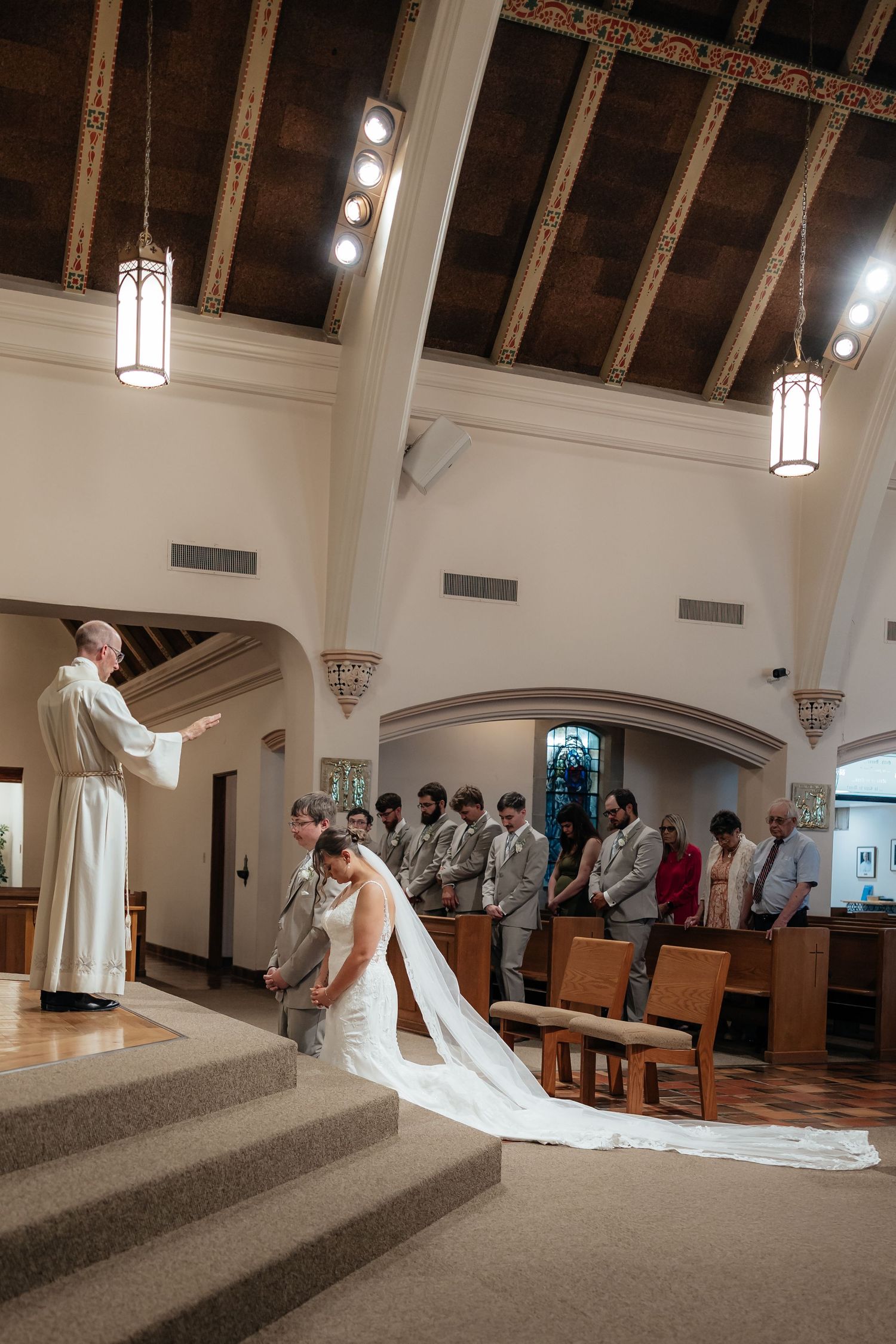 Western Nebraska Wedding Bride and groom kneel before officiant during ceremony in elegant church with wooden ceiling and pendant lights.