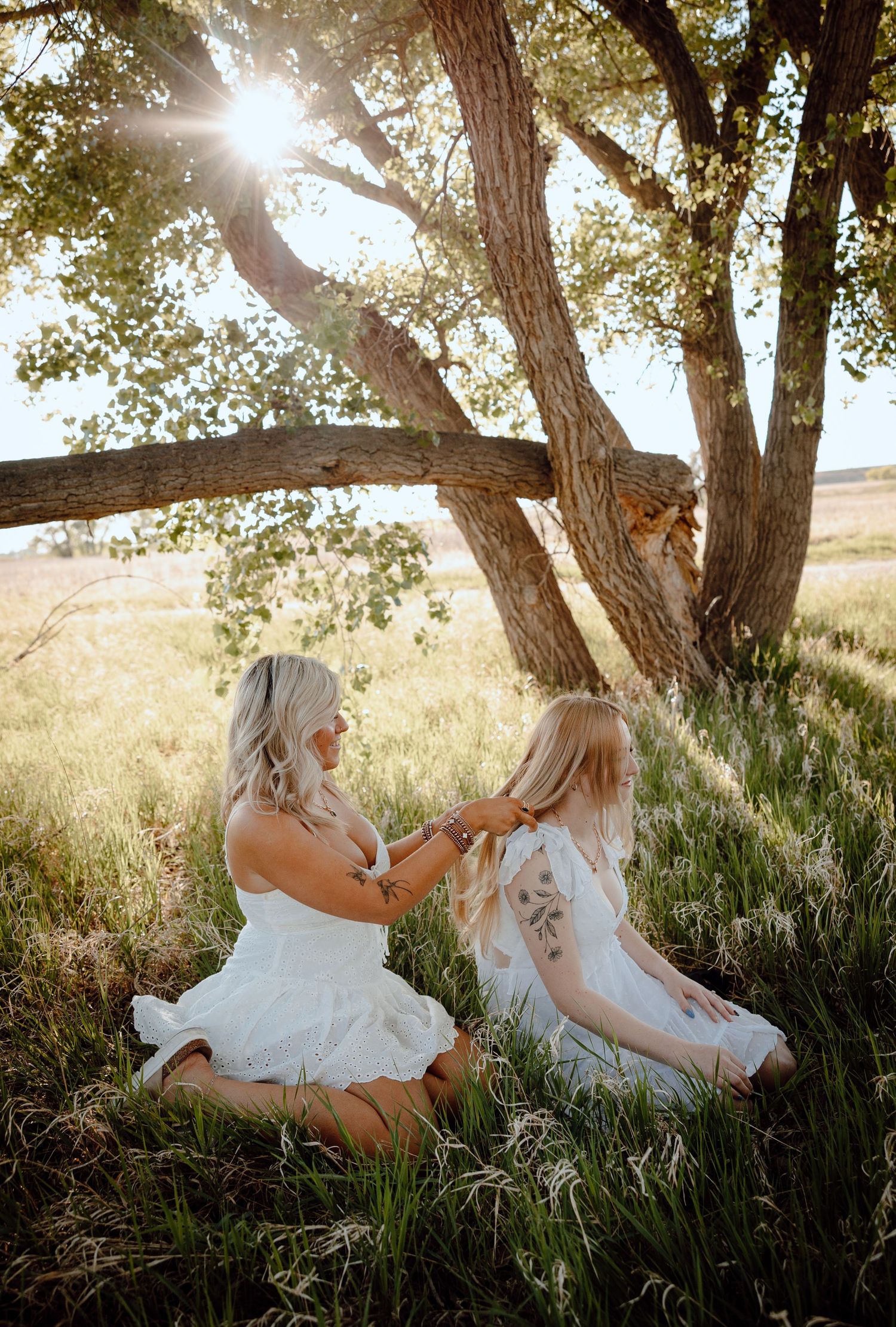 Hershey Nebraska Lisestyle Session two young girls sit in green grass braiding their hair with a golden sun behind them