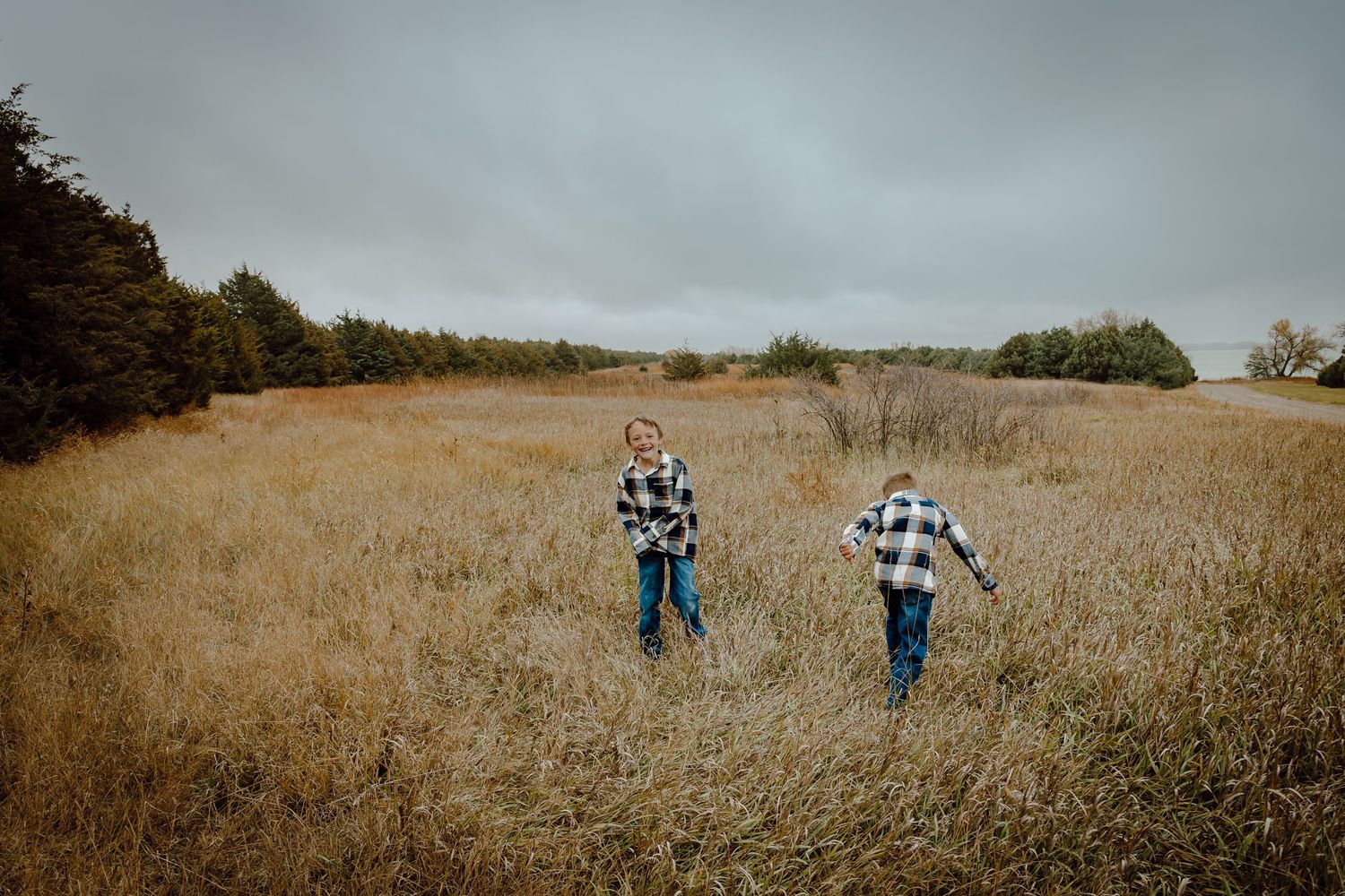 Sutherland Nebraska Family Session boys in plaid shirts run through a golden autumn field under overcast skies near a tree line.