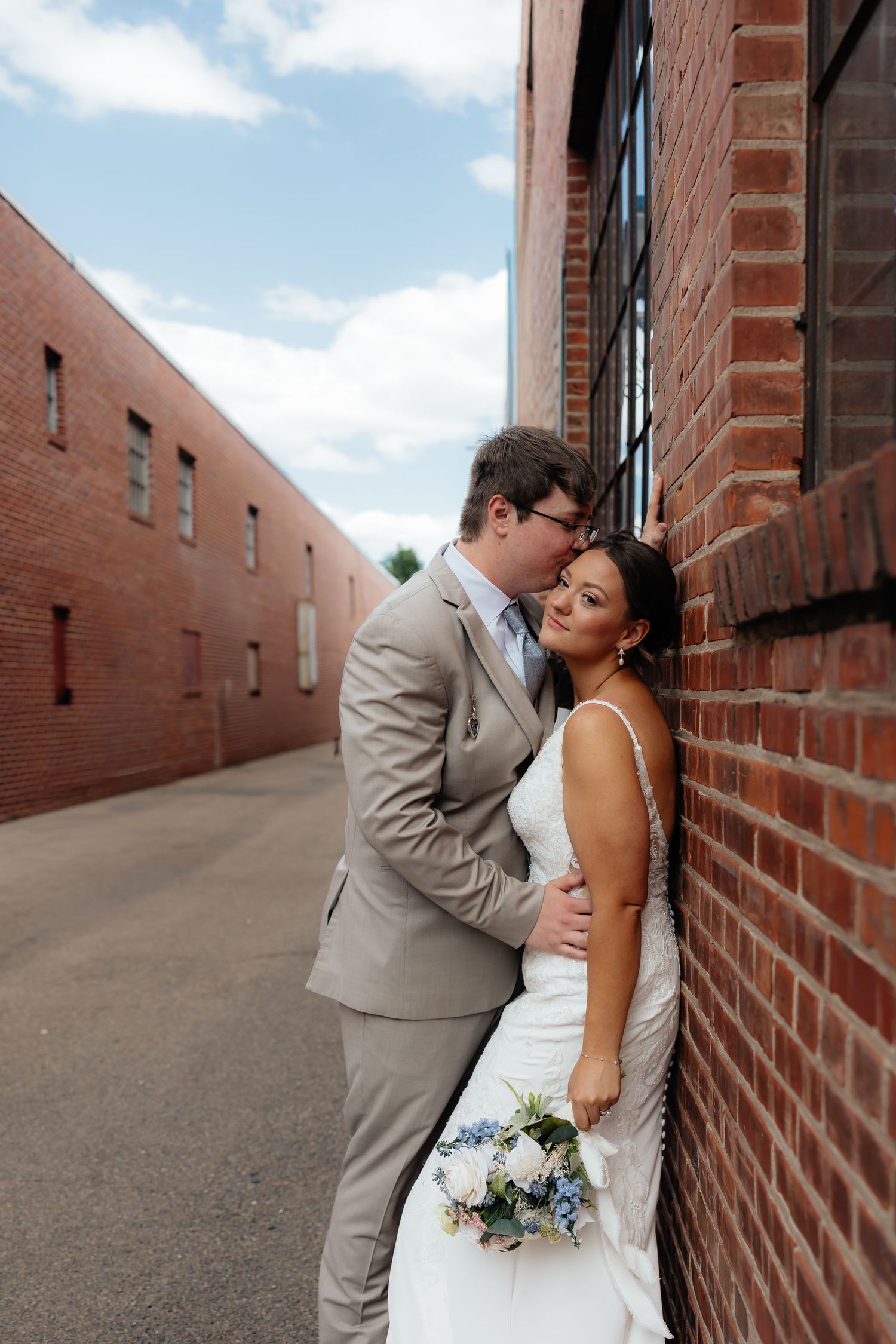 Western Nebraska Wedding Bride in white gown holds blue bouquet as groom in tan suit kisses her cheek in a brick alley.