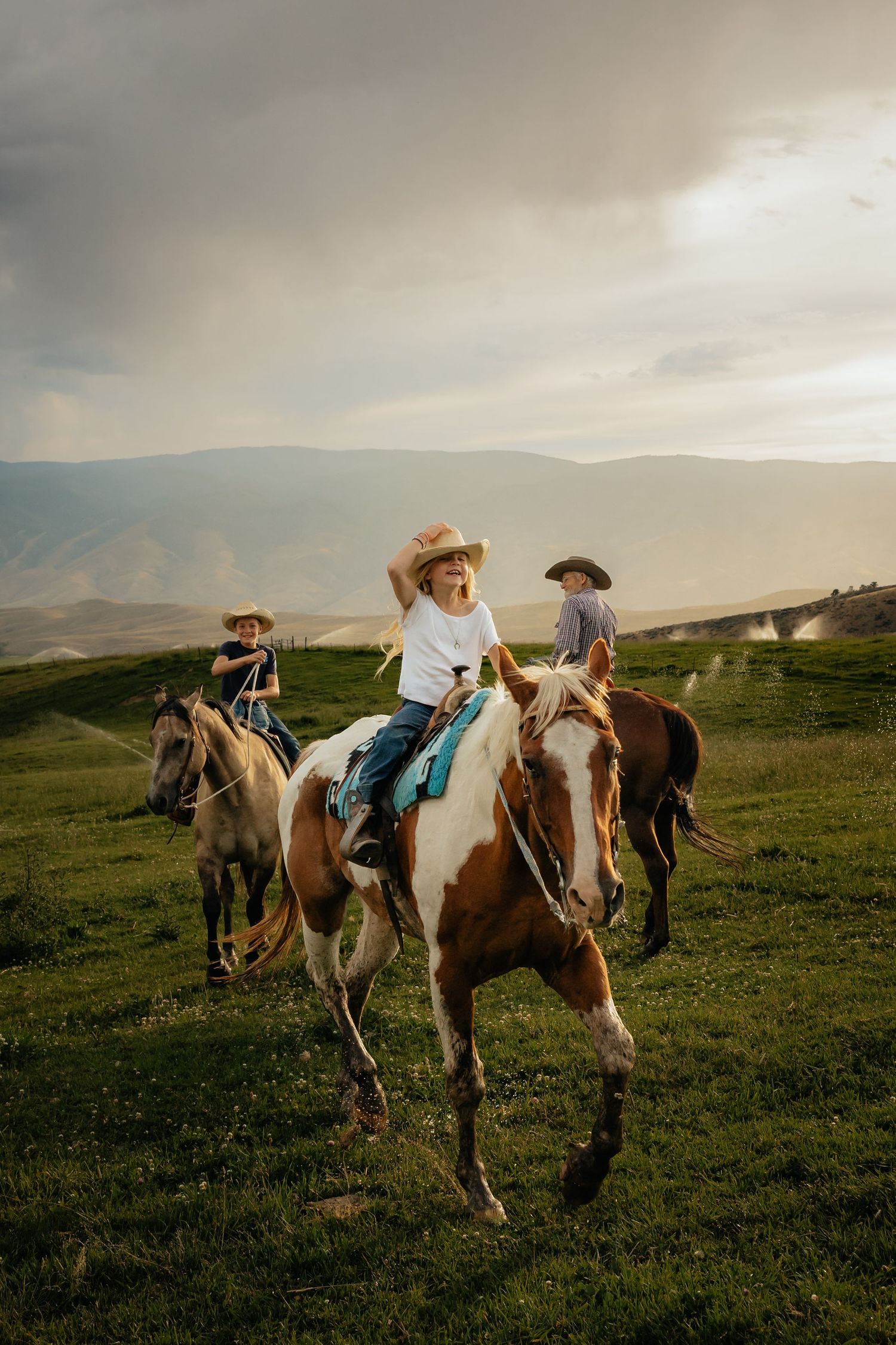 Western Nebraska Family Photography three children in cowboy hats ride horses across a lush green hillside
