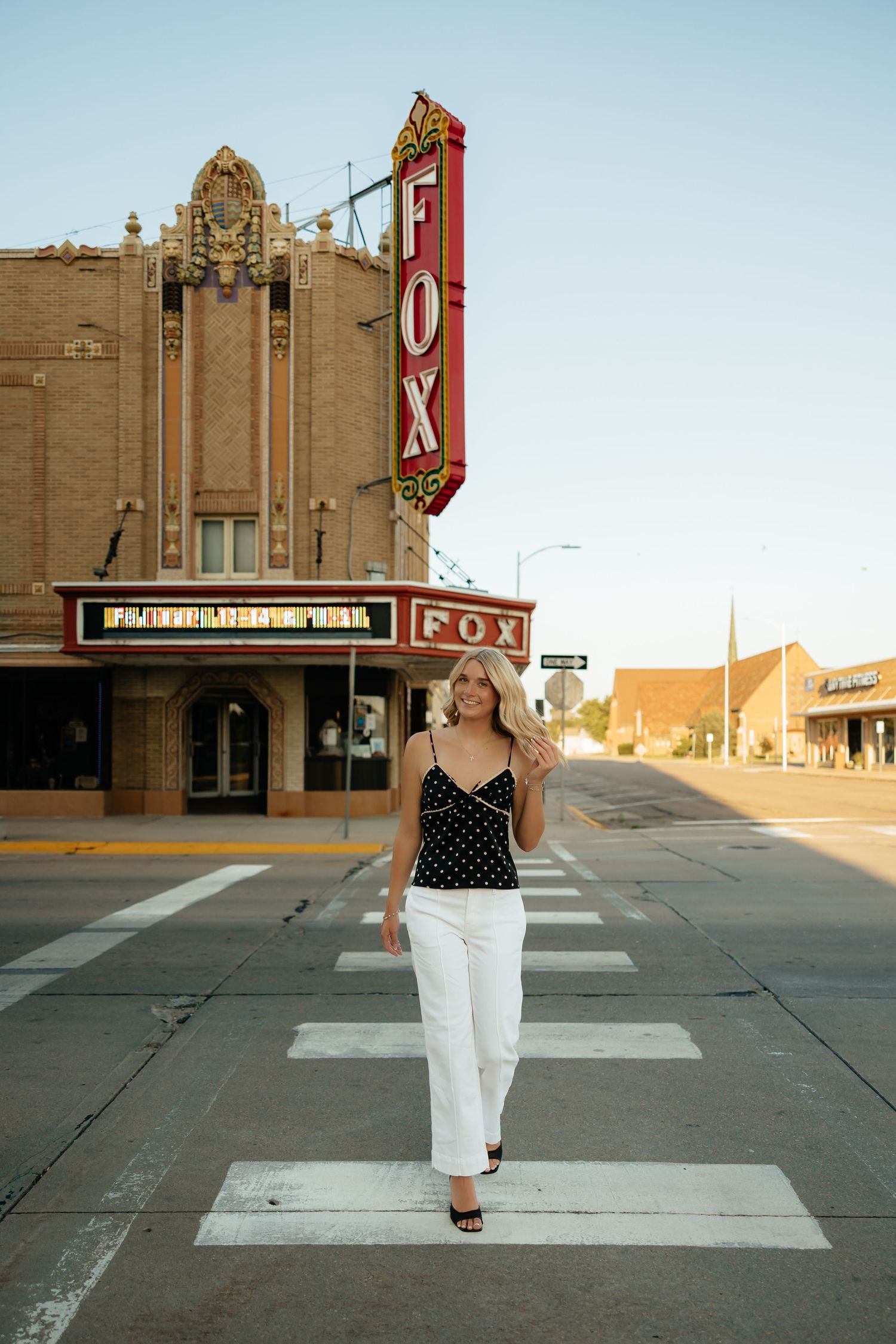 North Platte Nebraska Senior Session girl in white pants and polka dot top walking a crosswalk in front of the historic Fox Theatre.
