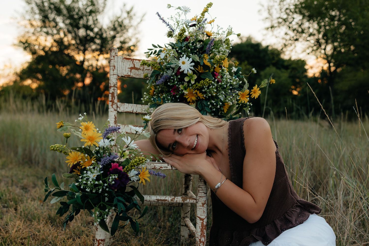 North Platte Nebraska Senior Session girl resting on a rustic white chair surrounded by wildflower bouquets in a golden field at sunset.