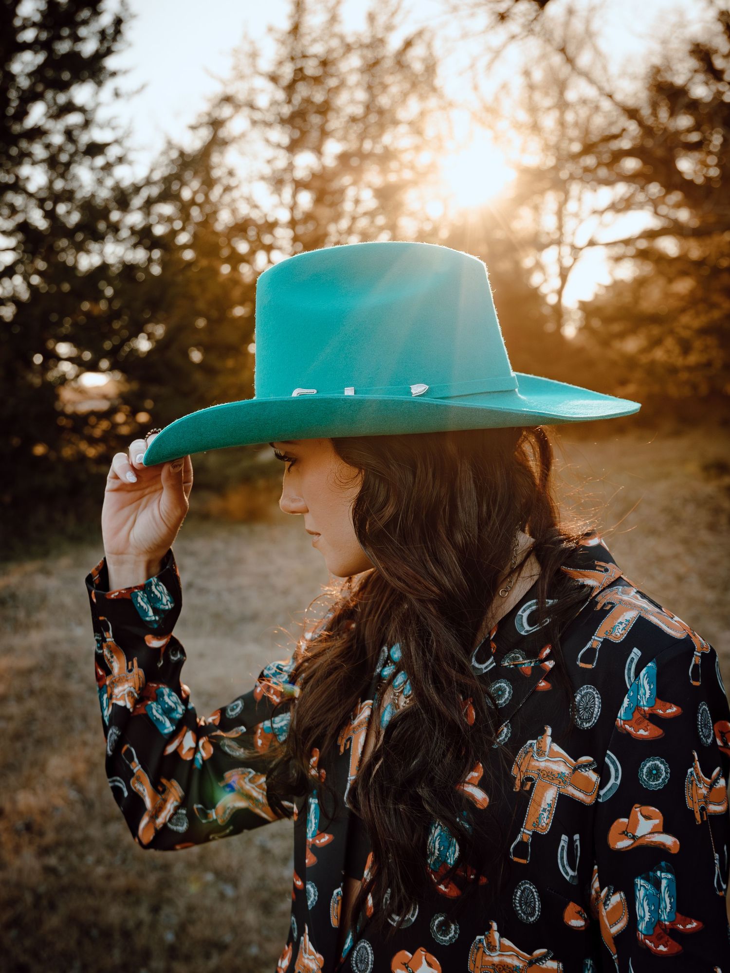 Western Nebraska Brand Session woman in turquoise wide-brim hat and western print shirt poses outdoors at golden hour sunset.