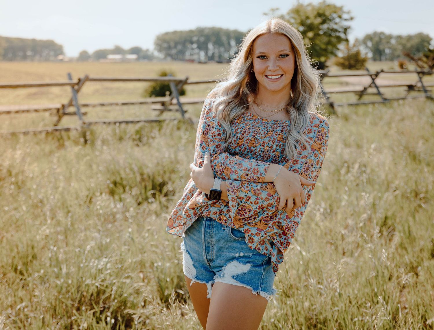 Chappell Nebraska Senior Session girl in floral top and denim shorts standing in a sunny field with a wooden fence backdrop.