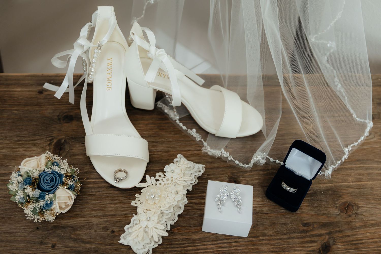 Western Nebraska Wedding accessories flat lay featuring white heels, veil, garter, bouquet, earrings, and ring box on wood.