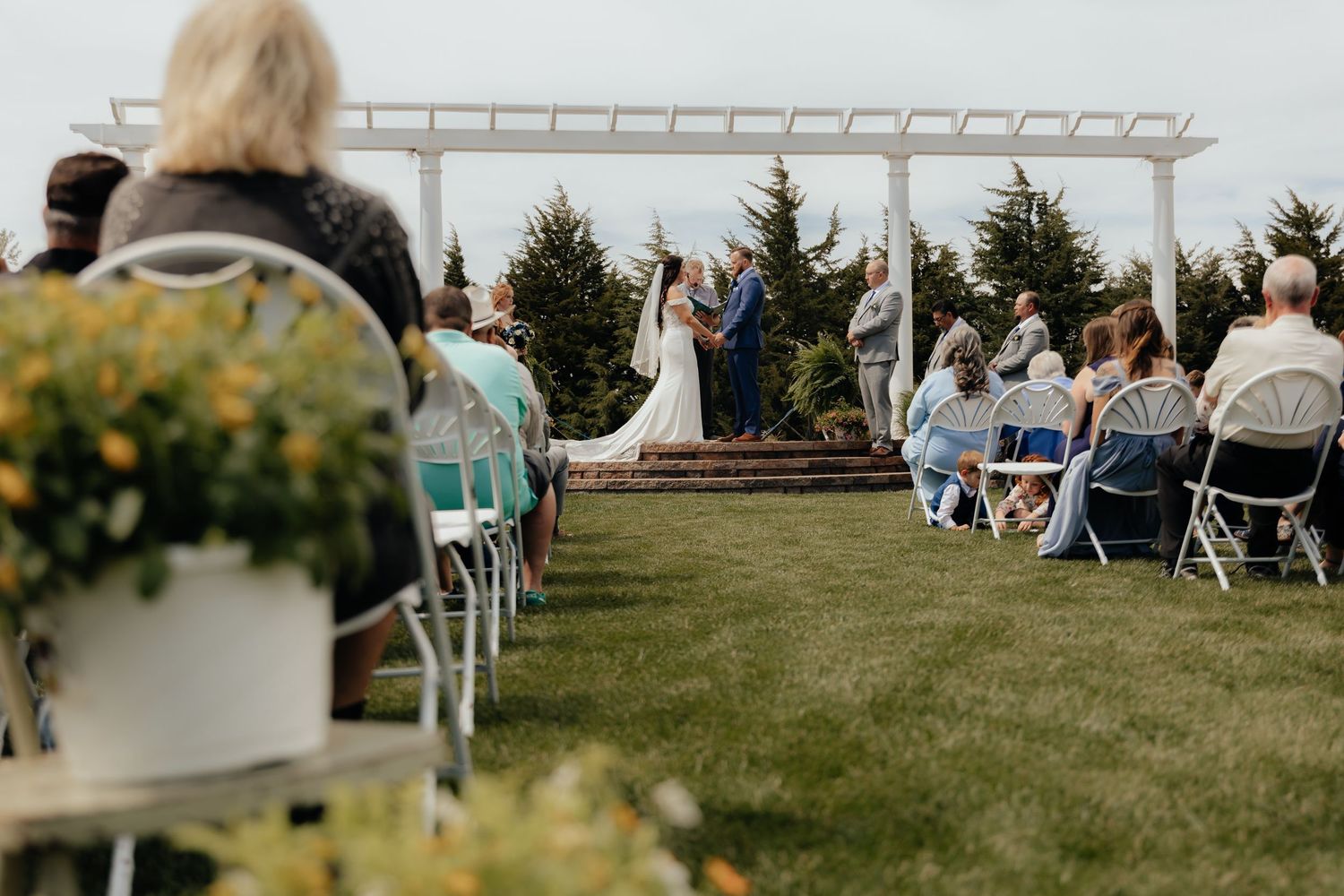 Western Nebraska wedding ceremony on green lawn, seated guests in folding chairs, and evergreen trees backdrop.