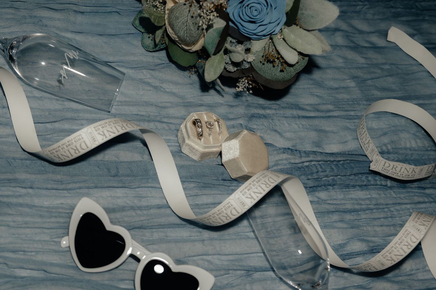 Western Nebraska Wedding flat lay with heart sunglasses, ribbon, ring box, champagne glasses, and floral bouquet
