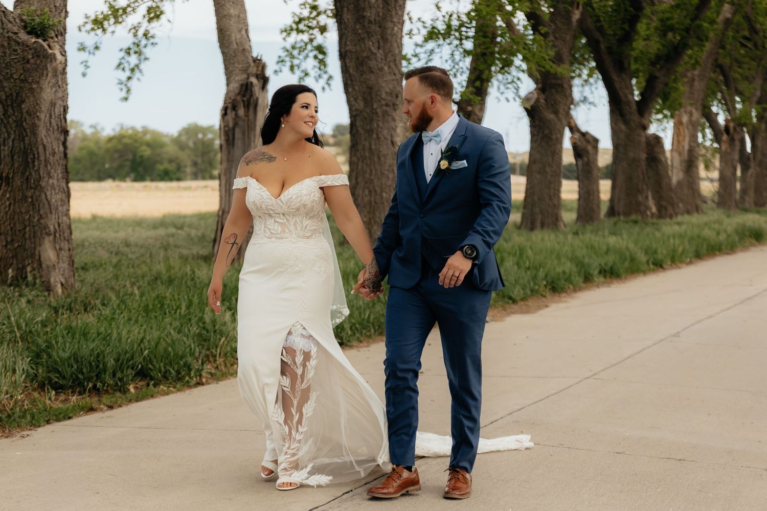 Western Nebraska Wedding Bride in white off-shoulder gown and groom in navy suit walk hand-in-hand on a tree-lined path.