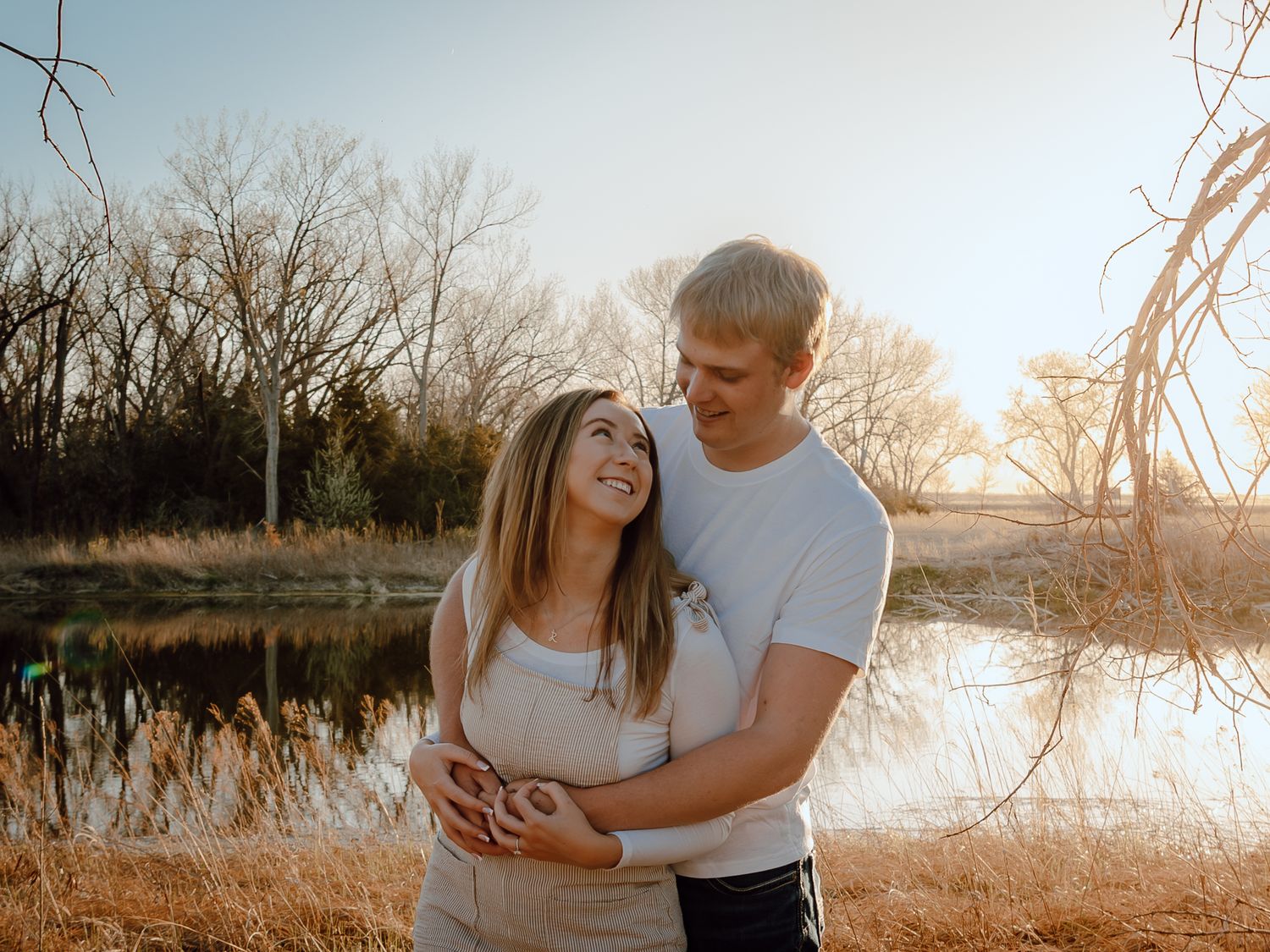 Paxton Nebraska Engagement Session couple embraces and smiles by a sunlit lake surrounded by bare trees