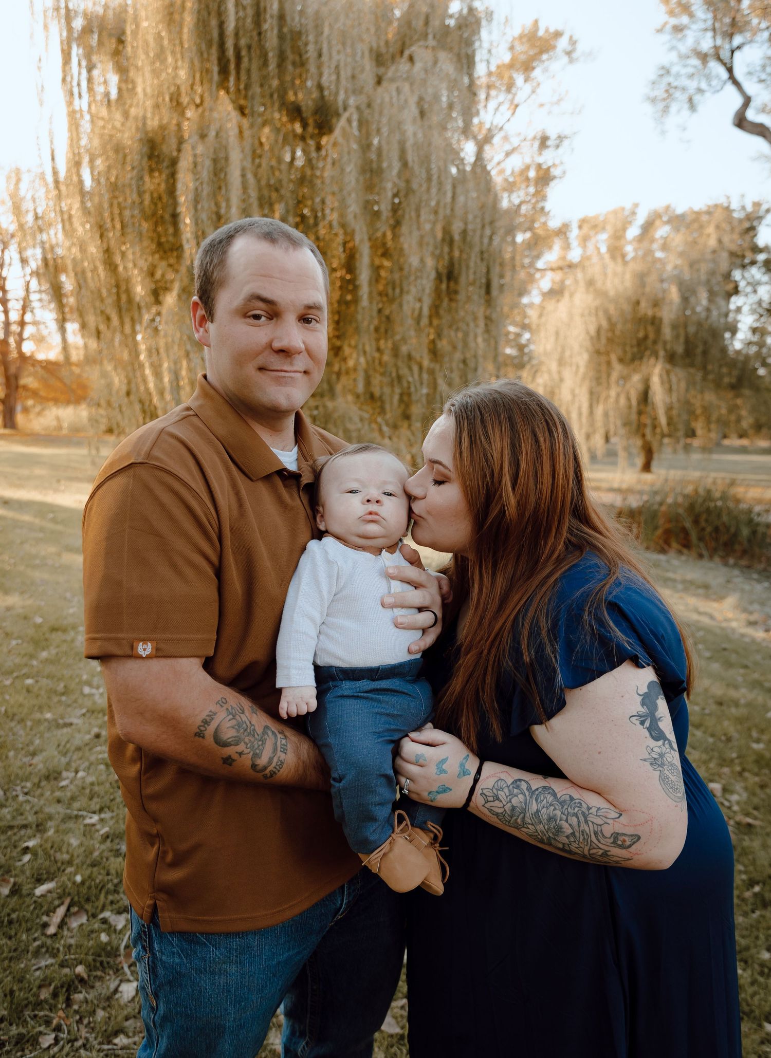 North Platte Nebraska Family Session parents kissing their baby near a willow tree in warm golden light.