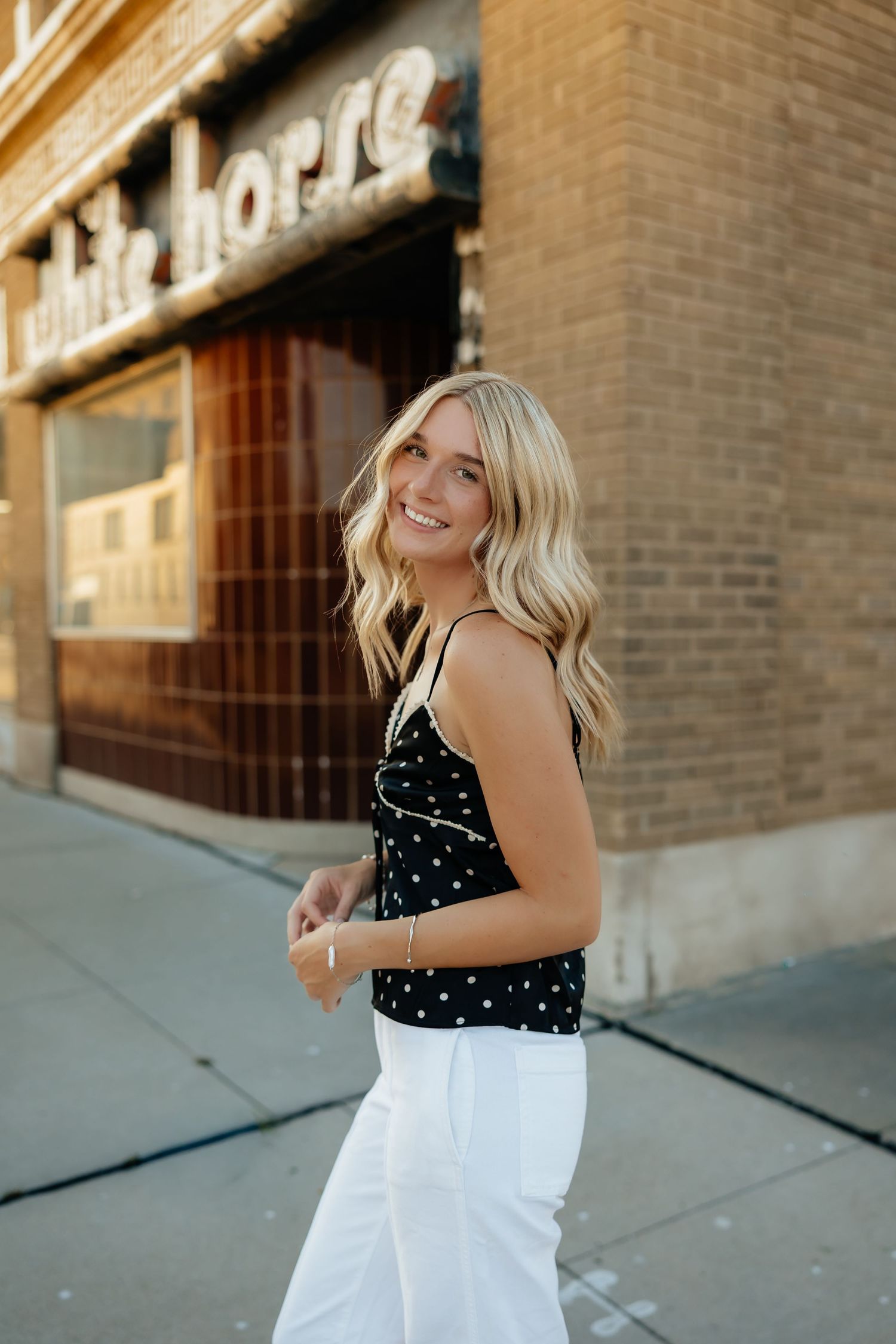 North Platte Nebraska Senior Session girl in black polka dot top and white pants standing on sidewalk outside White Horse brick building.