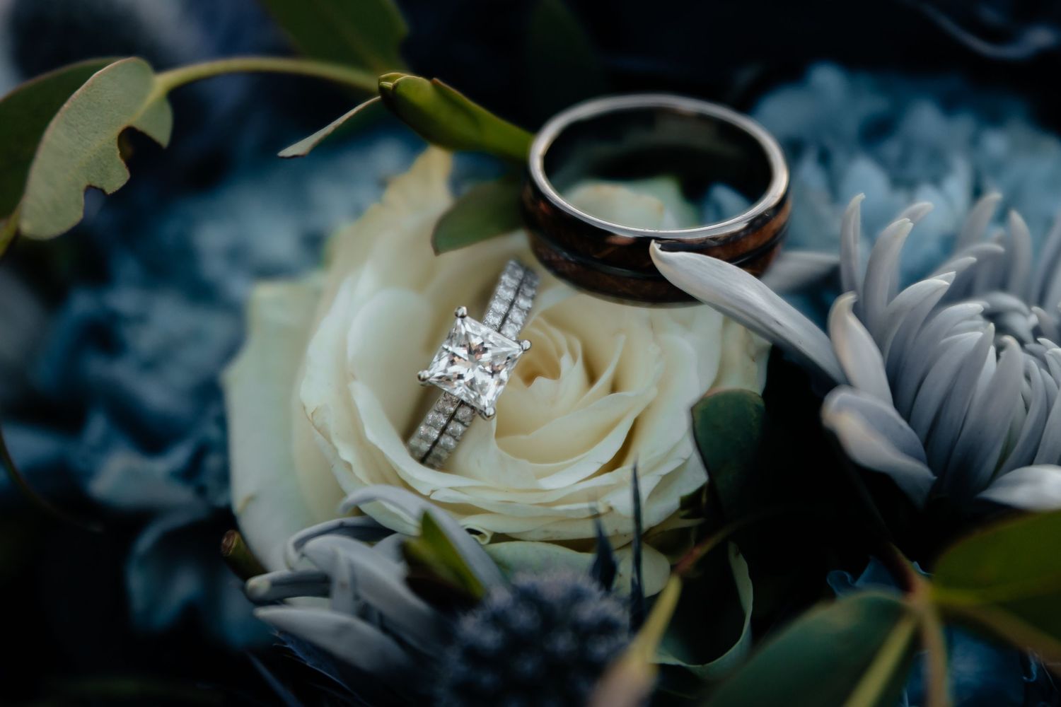 Western Nebraska Wedding rings resting on white and blue flowers, featuring a diamond engagement ring and a dark wood band.
