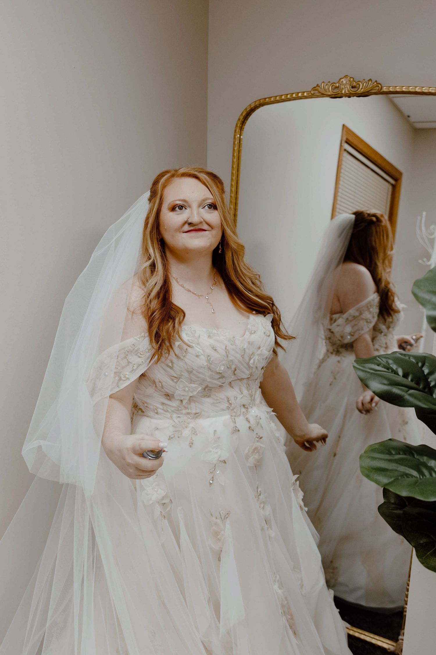 Western Nebraska Wedding Photography a bride in floral lace off-shoulder wedding gown with veil stands before gold-framed mirror.