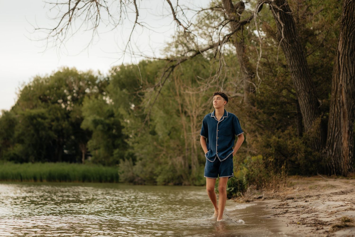 North Platte Nebraska Senior Session young man walking barefoot along a sandy shoreline with lush green trees in the background.