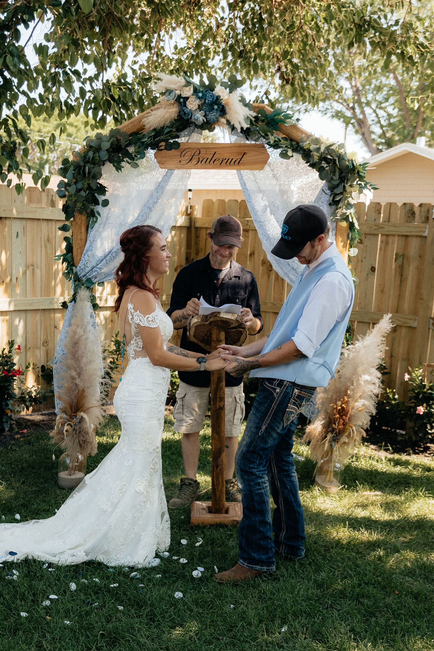 Western Nebraska Wedding backyard ceremony with bride in white gown and groom in jeans under floral arch with greenery.