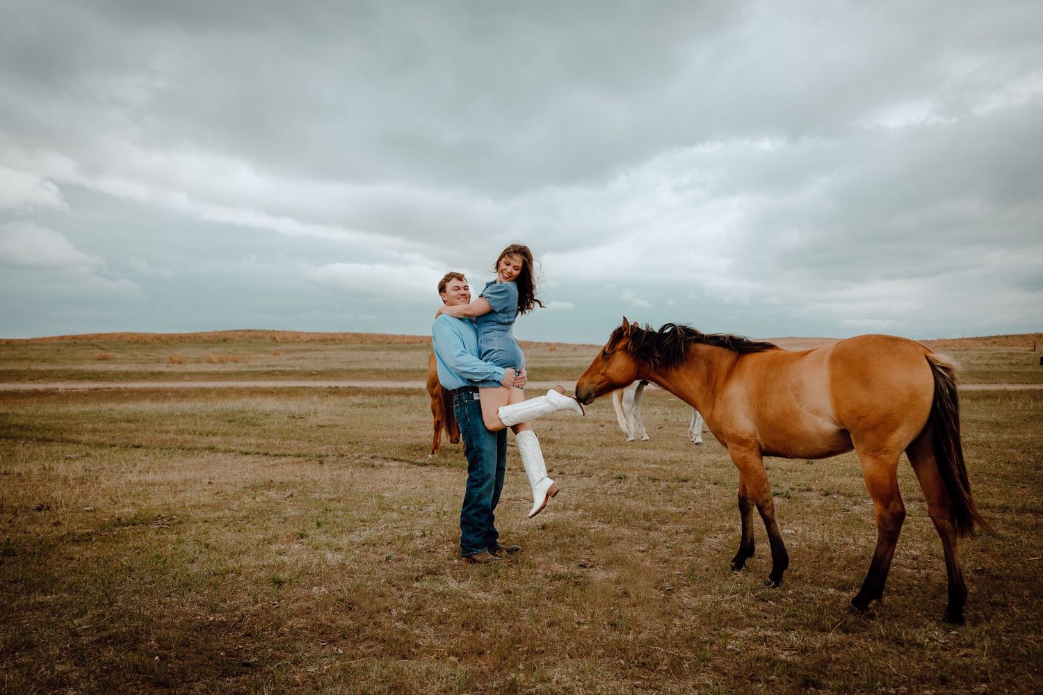 Nebraska Sandhills Engagement Session Couple playfully posing with a brown horse in an open field under a cloudy sky.