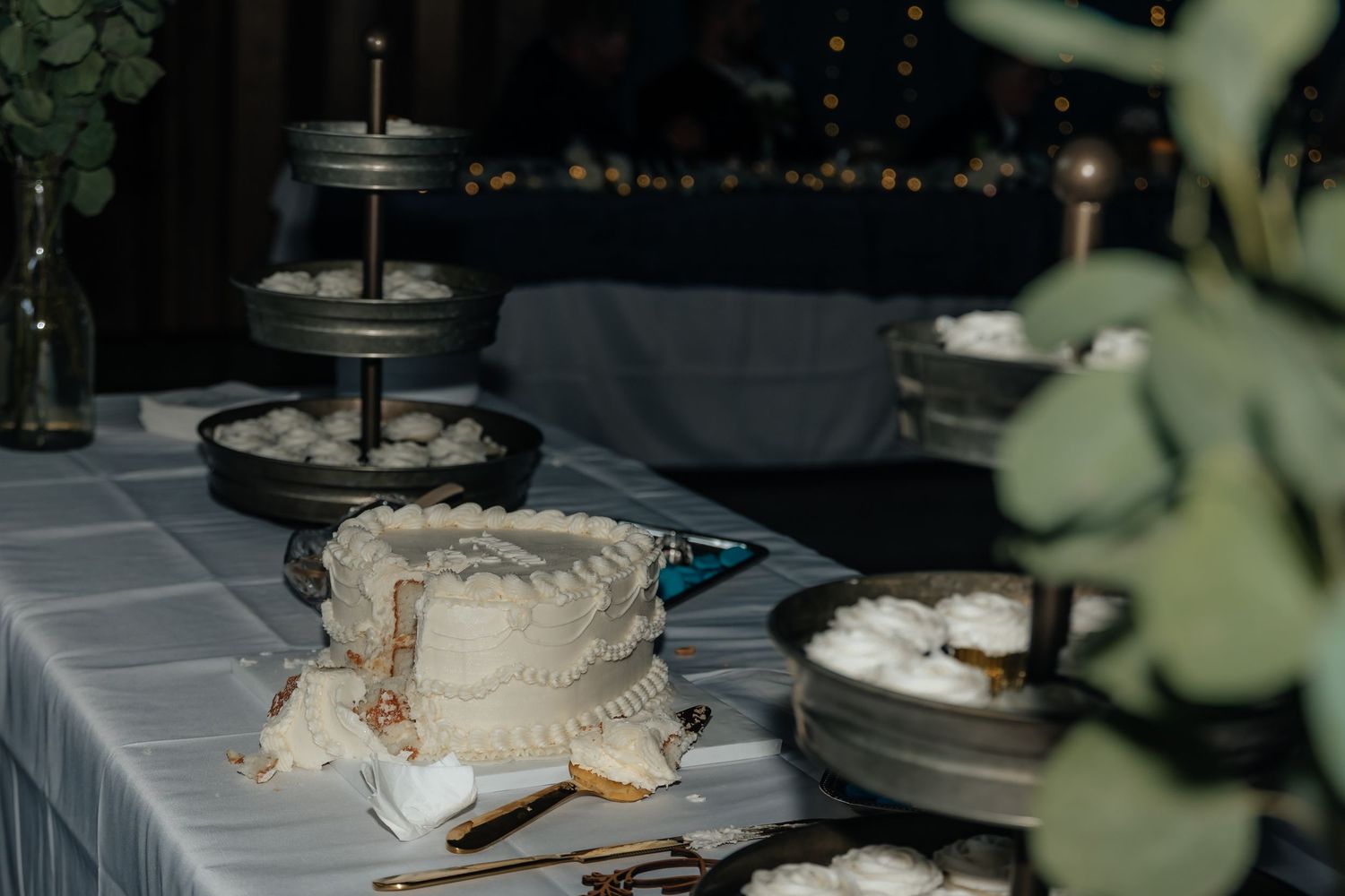 Western Nebraska Wedding Elegant white cake on a table with tiered stands and greenery in a dimly lit reception venue.
