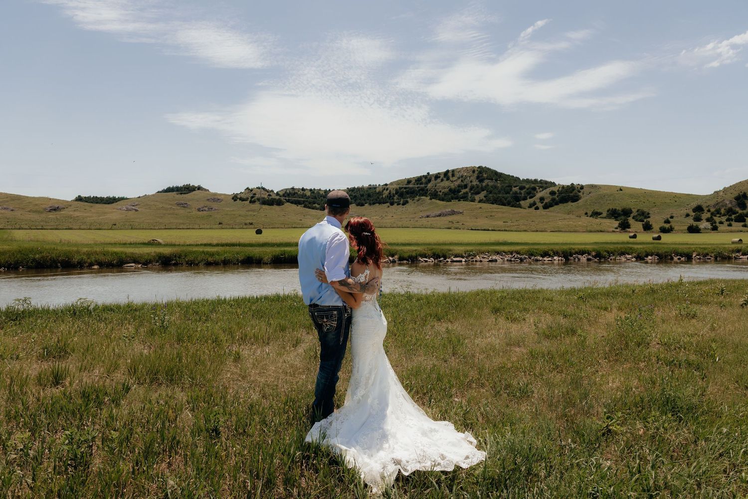 Western Nebraska Wedding Bride and groom embrace in a lush green field with a river and rolling hills backdrop on their wedding day.