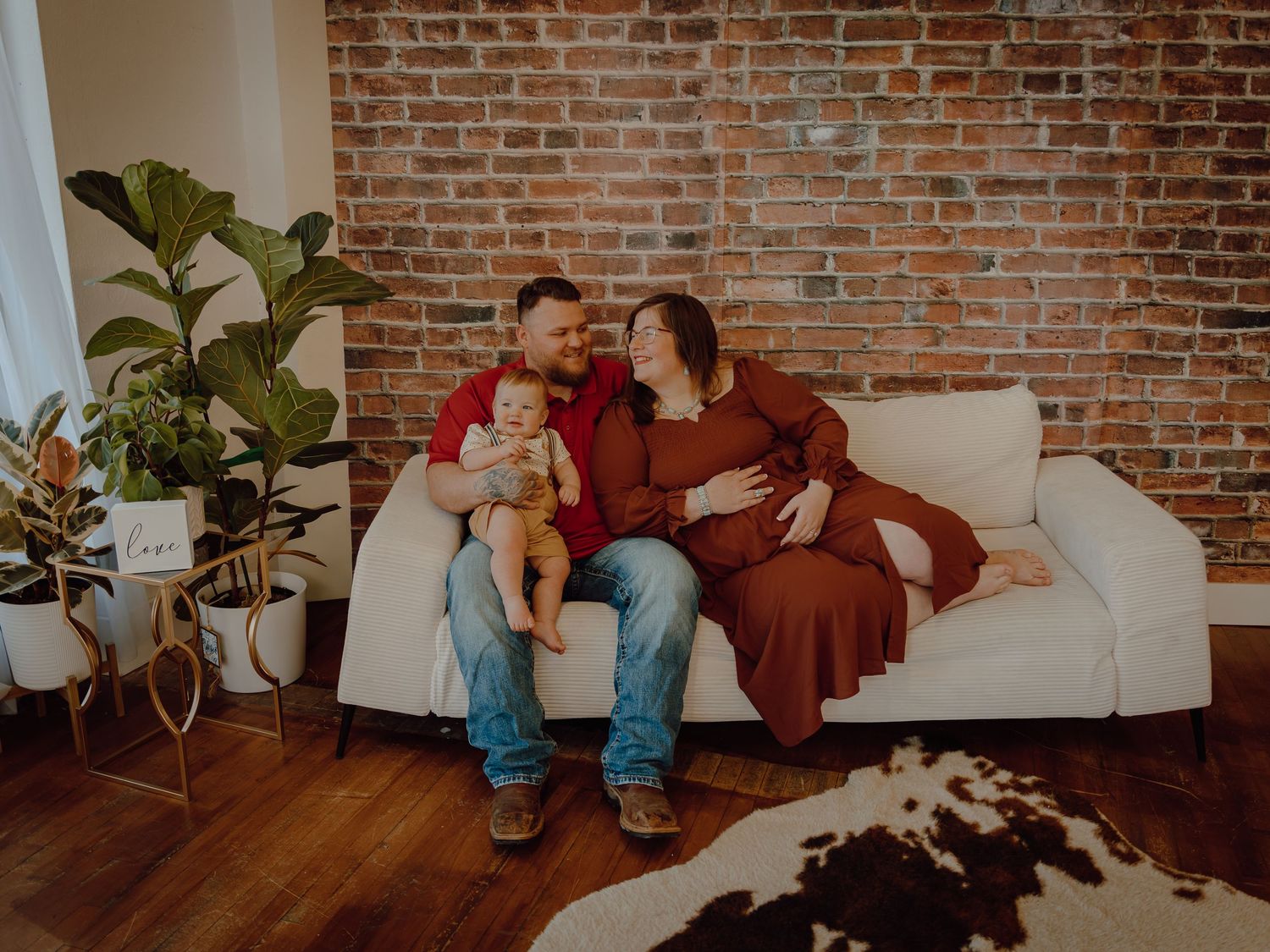 Western Nebraska Family Session couple with baby sitting on white couch against brick wall in a cozy studio with hardwood floors.
