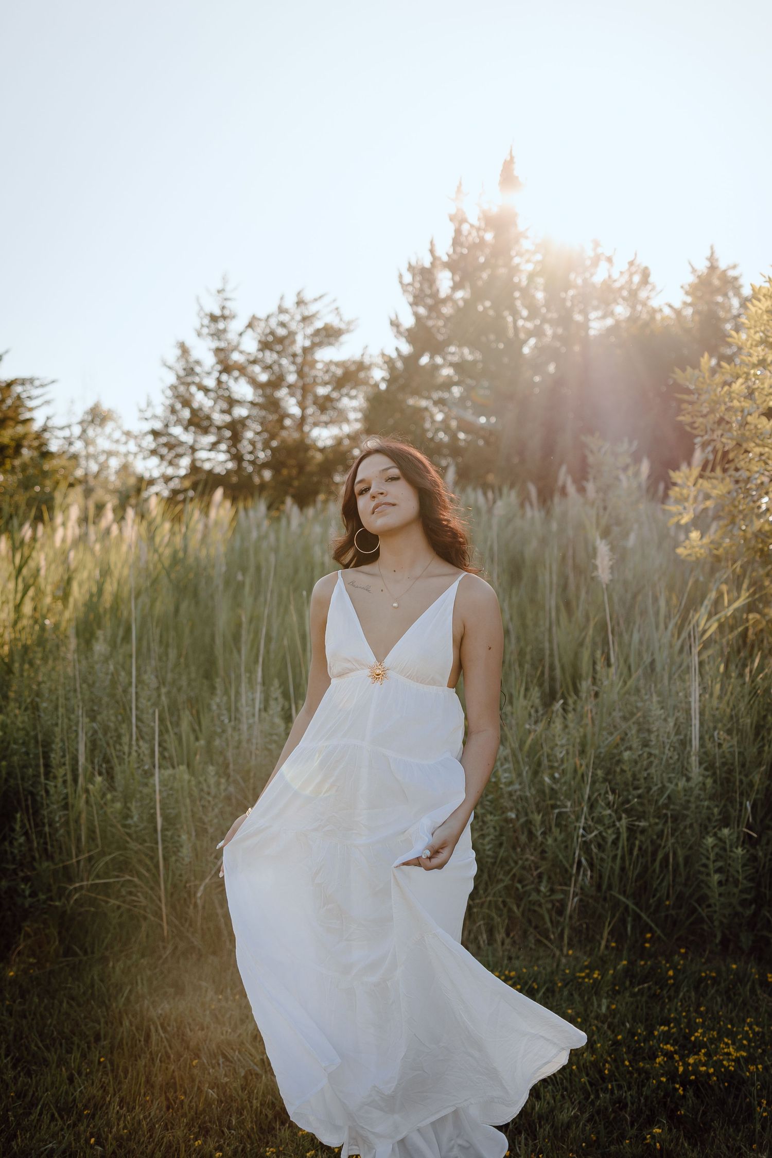 western nebraska senior session girl in flowing white dress standing in golden sunlight among tall grasses and trees.