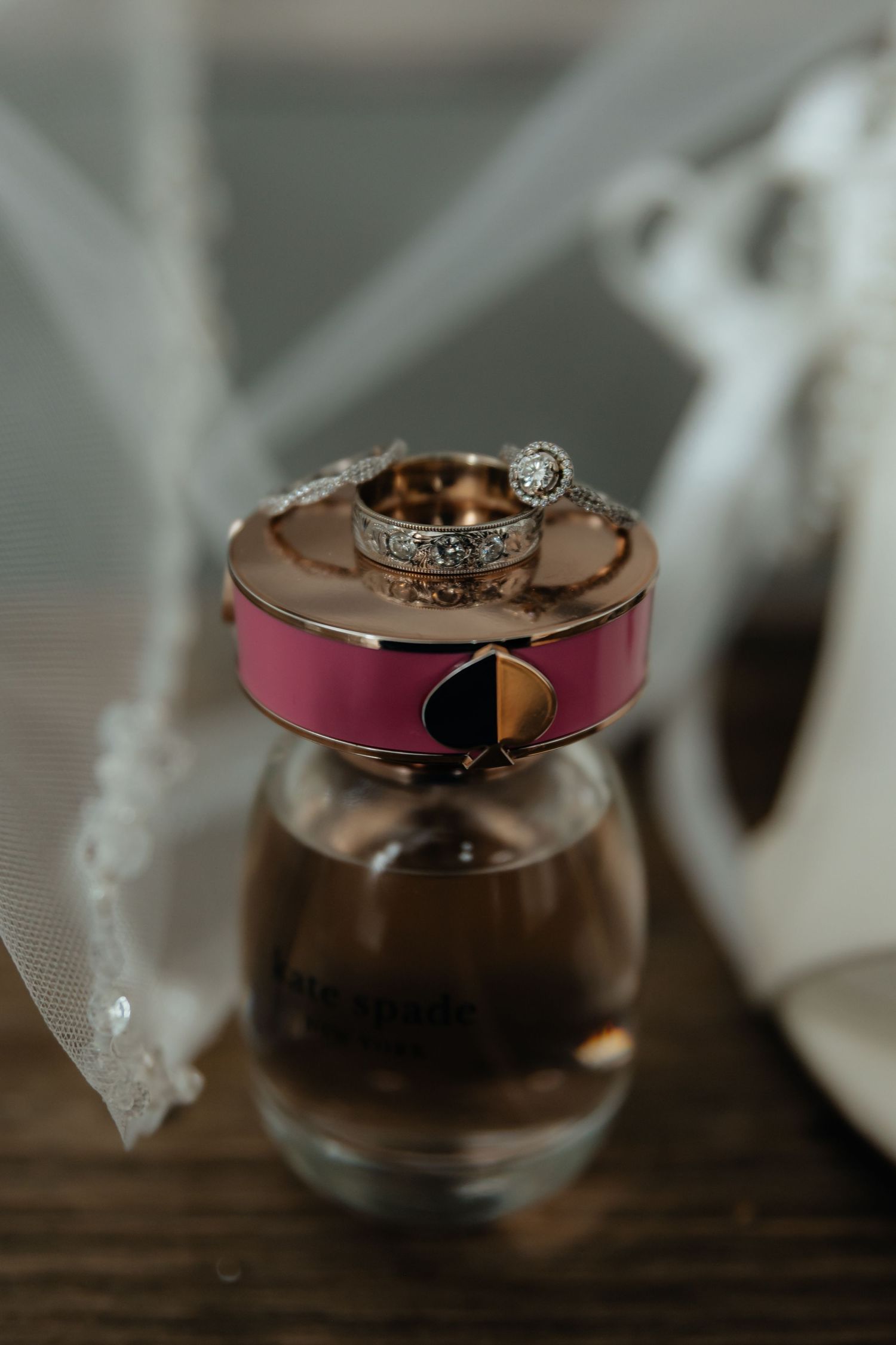 Western Nebraska Wedding rings and bands resting on a perfume bottle, with a bridal veil and white heels in the background.