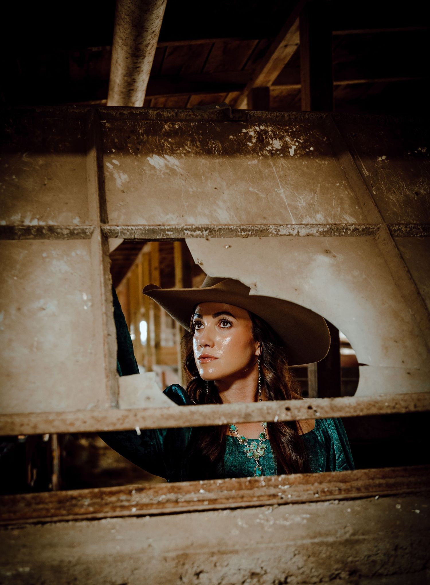 a north platte nebraska brand session cowgirl in teal shirt and wide-brim cowboy hat gazes upward through rustic wooden barn frame