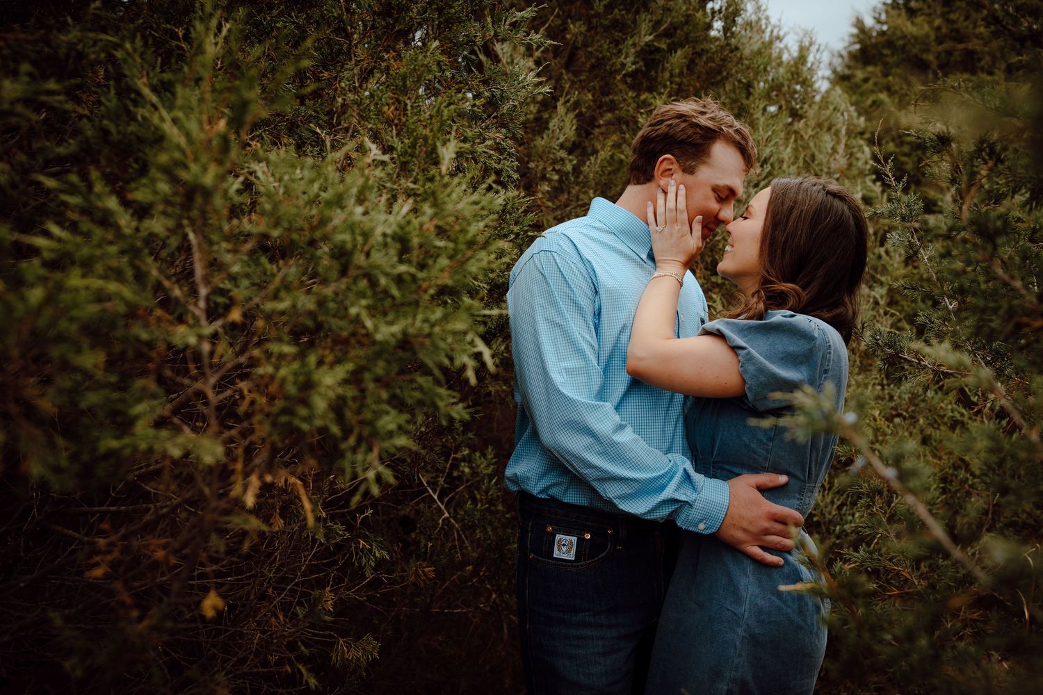 Nebraska Sandhills Engagement Session Couple sharing a romantic kiss surrounded by lush green evergreen trees