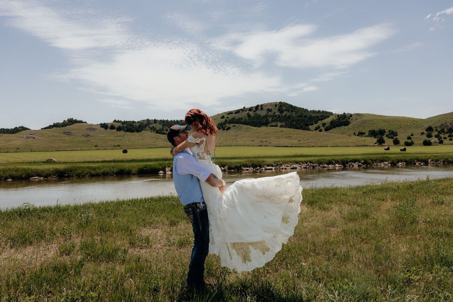 western Nebraska wedding photography Groom lifting bride in white dress by a river with green hills under blue sky.