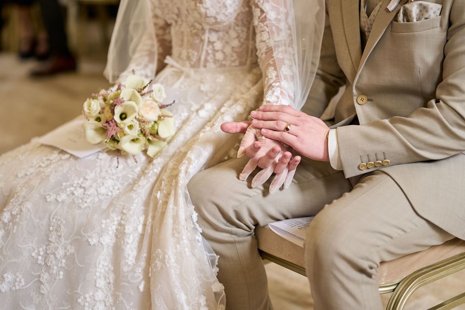 A menyasszony és a vőlegény egymást fogó kezei közelről az esküvői szertartáson, mellettük elegáns virágcsokorral.