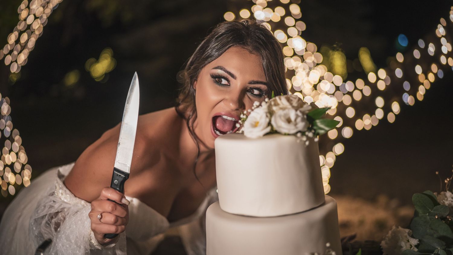 Pánt nélküli ruhás menyasszony játékosan tartja a tortakést a fehér, emeletes esküvői torta felvágásakor a WeddingWoodban.