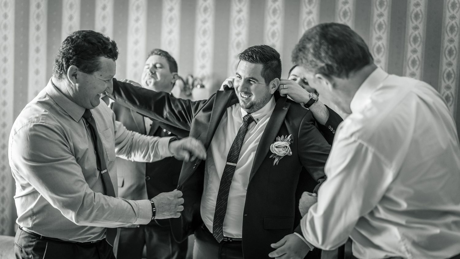 A vőlegény öltönyét igazító férfiak az esküvői készülődés közben, fekete-fehér felvételen.
