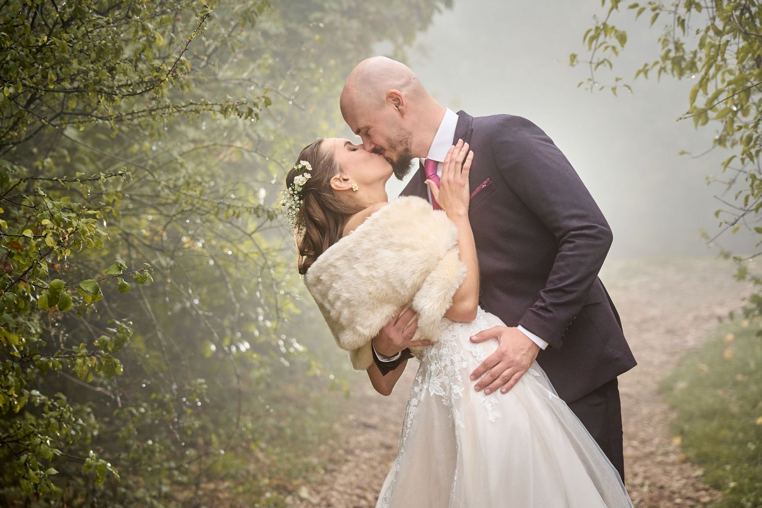 Bride and groom sharing a romantic dip kiss on a misty forest path, bride wearing a fur stole.