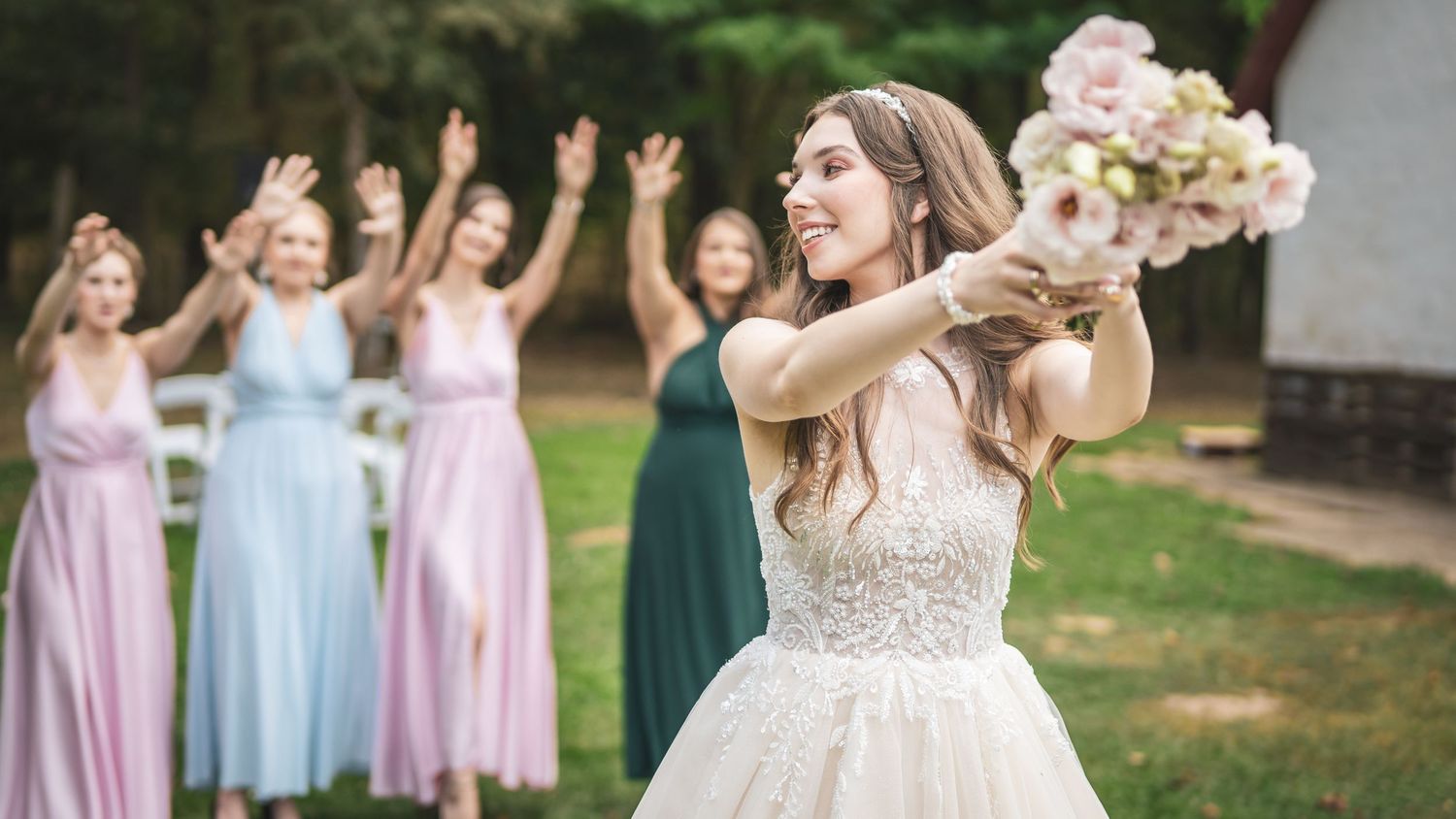 Csipkeruhás menyasszony rózsaszín rózsacsokrot dob a színes ruhás koszorúslányoknak a WeddingWood szabadtéri esküvői helyszínén.