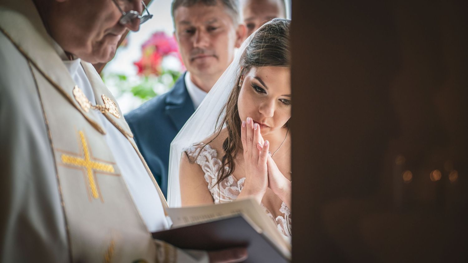 A menyasszony összetett kézzel áll a vőlegény mellett a templomi esküvőn, miközben a pap könyvből olvas.