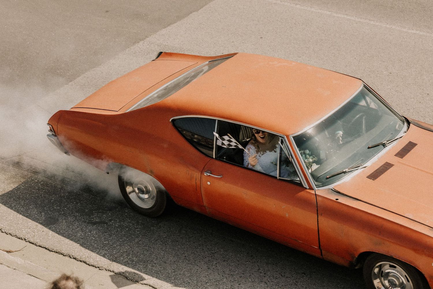 Vintage orange muscle car performing a burnout on a street, creating dramatic smoke from rear tires.