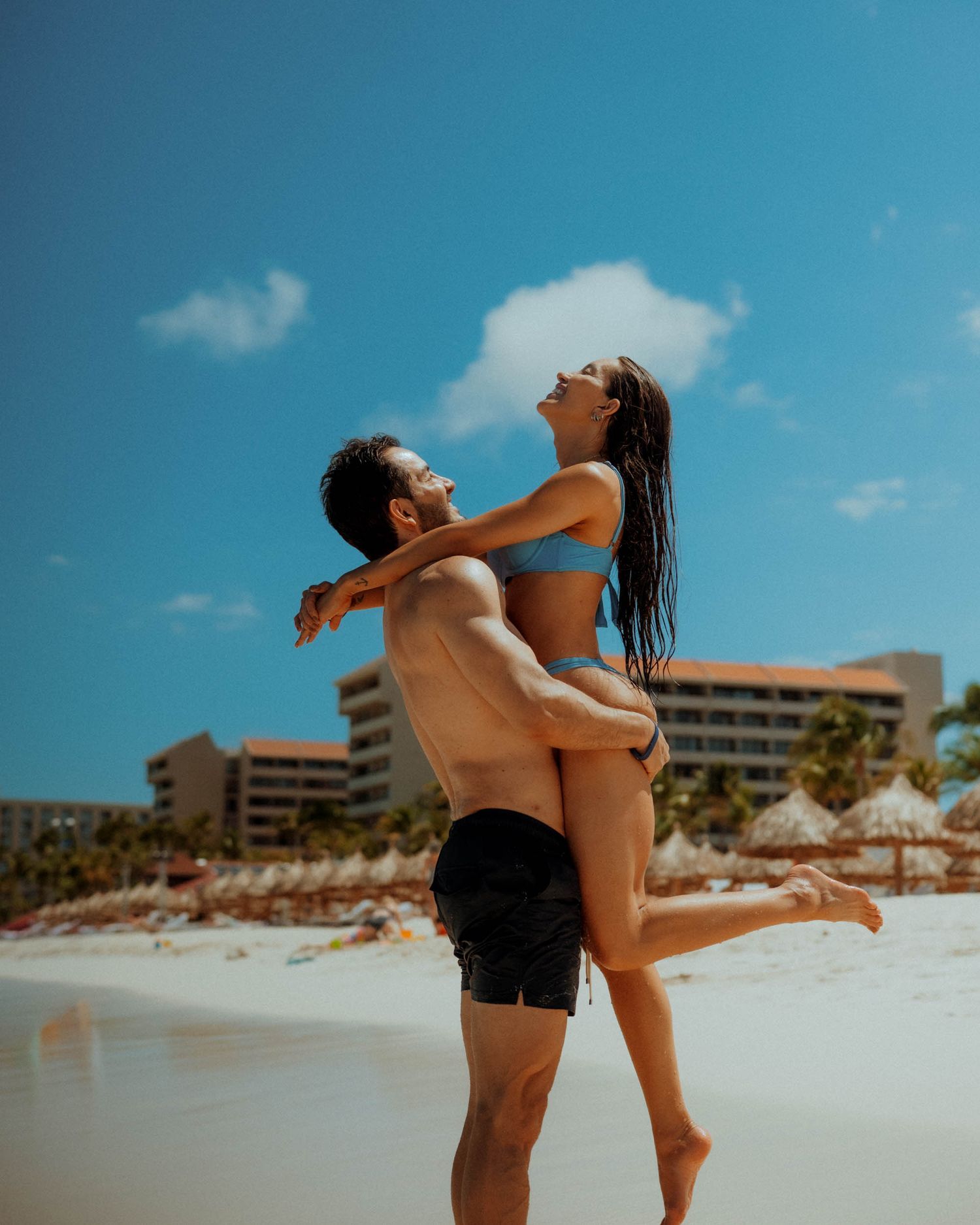 Man lifting woman in a blue bikini on a sunny beach with resort buildings visible in the background.