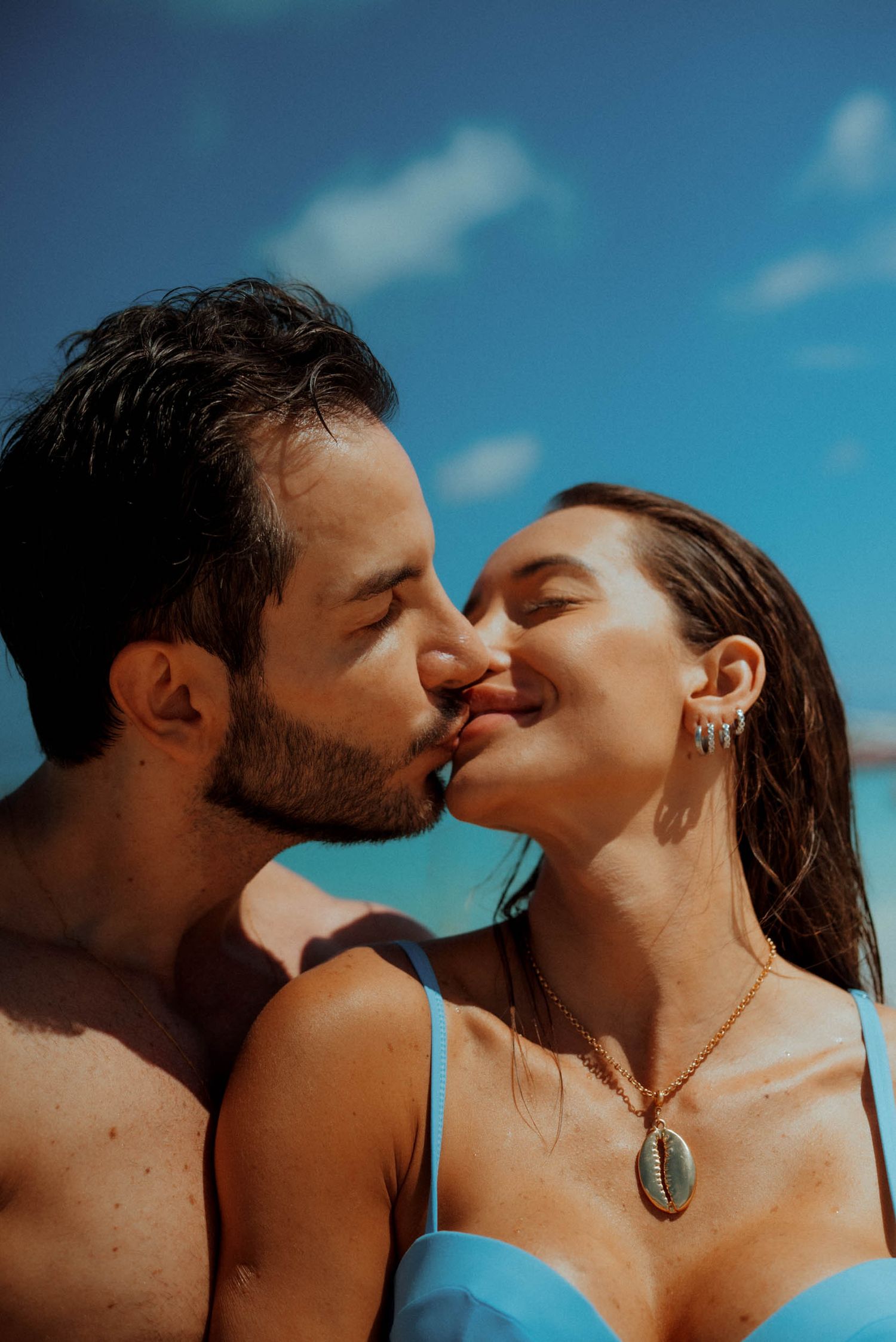 Couple sharing a romantic kiss on a sunny beach, woman wearing a blue bikini and shell necklace against blue sky.