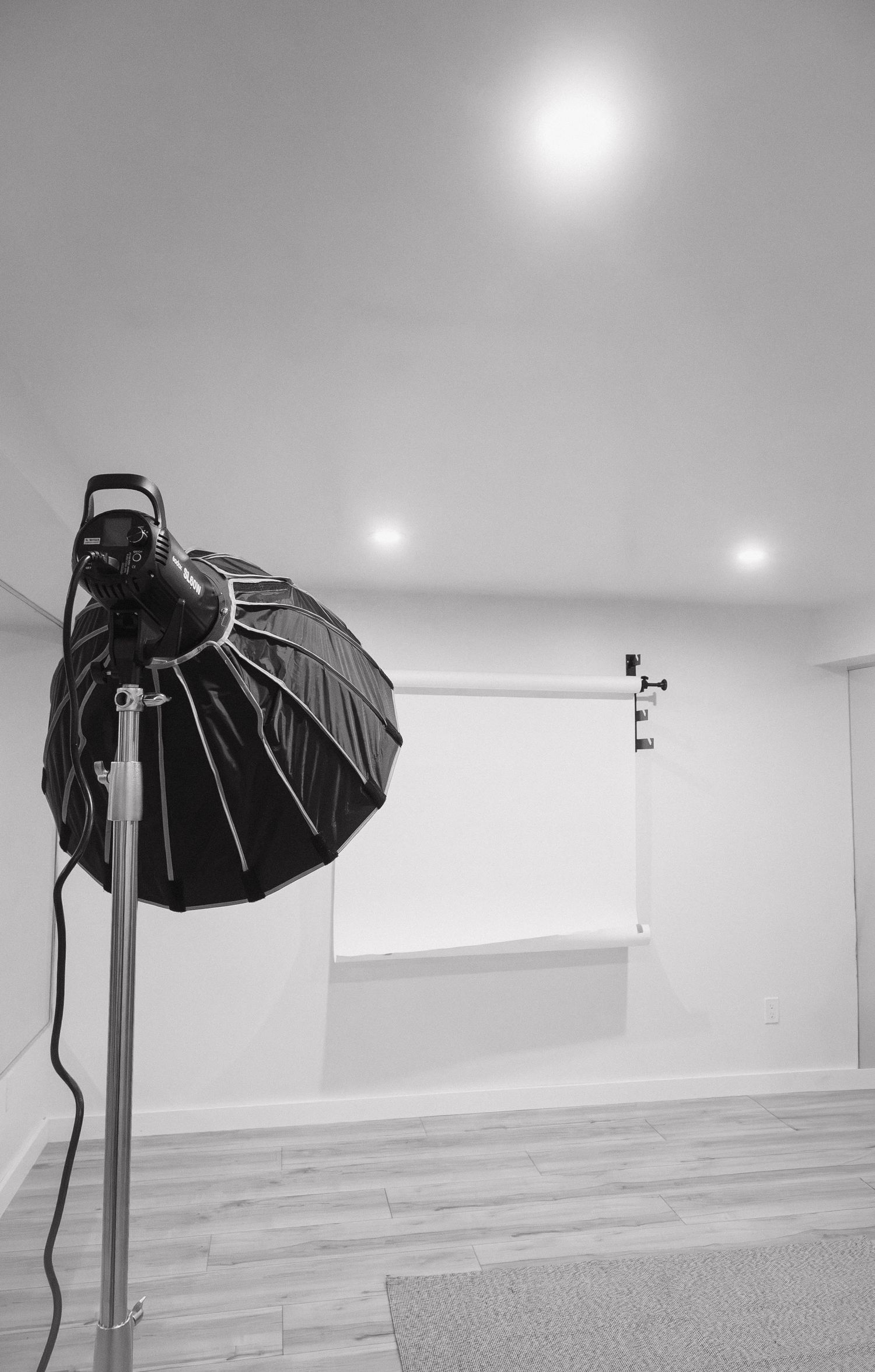 Black and white photo of a photography studio with a softbox light on a tripod and white backdrop.