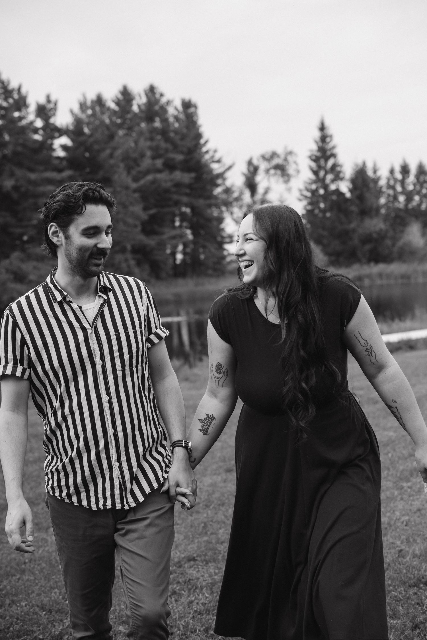 Black and white photo of a laughing couple holding hands and walking outdoors near trees and a pond.
