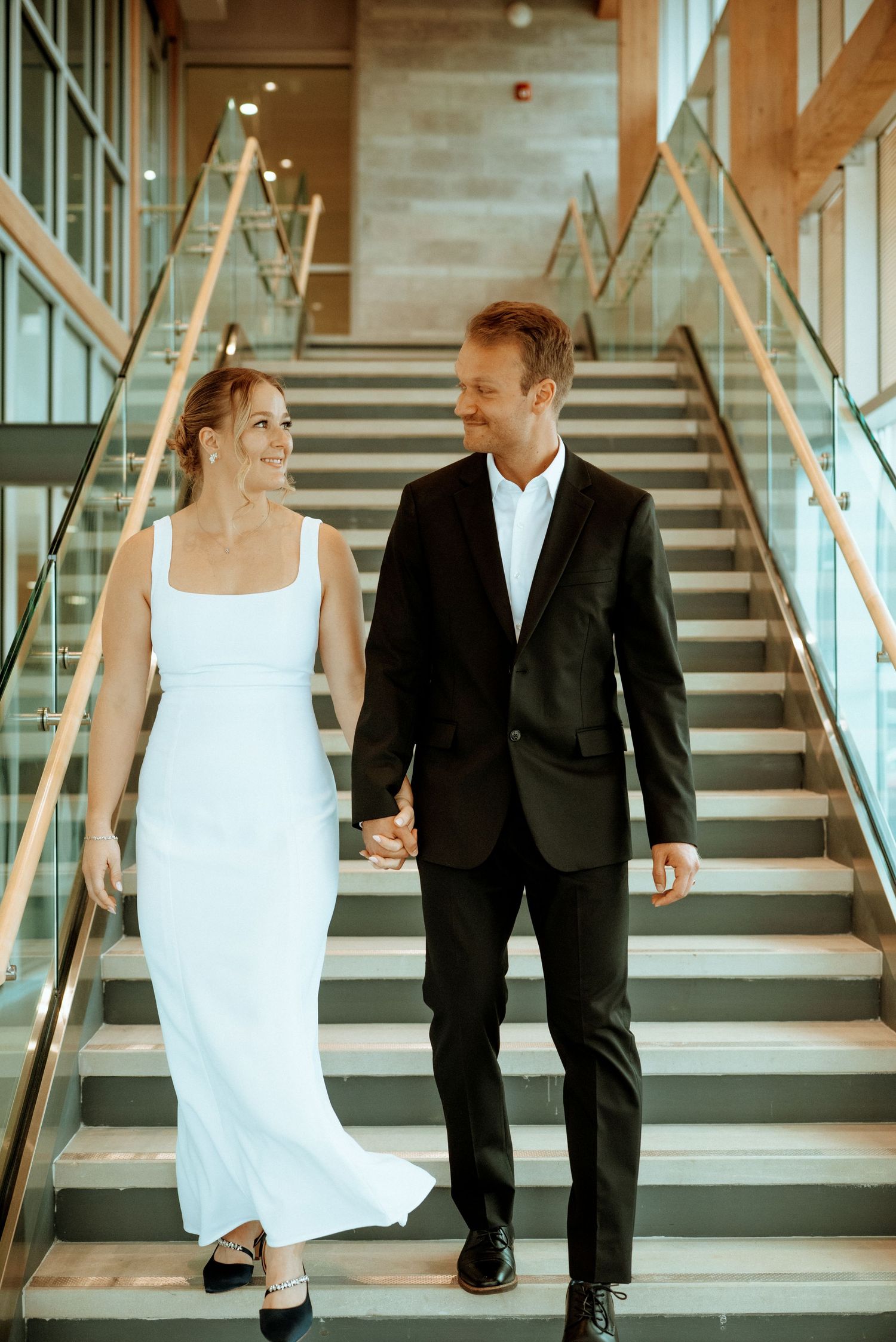 Bride in white gown and groom in black suit walk hand-in-hand down modern staircase, smiling at each other.