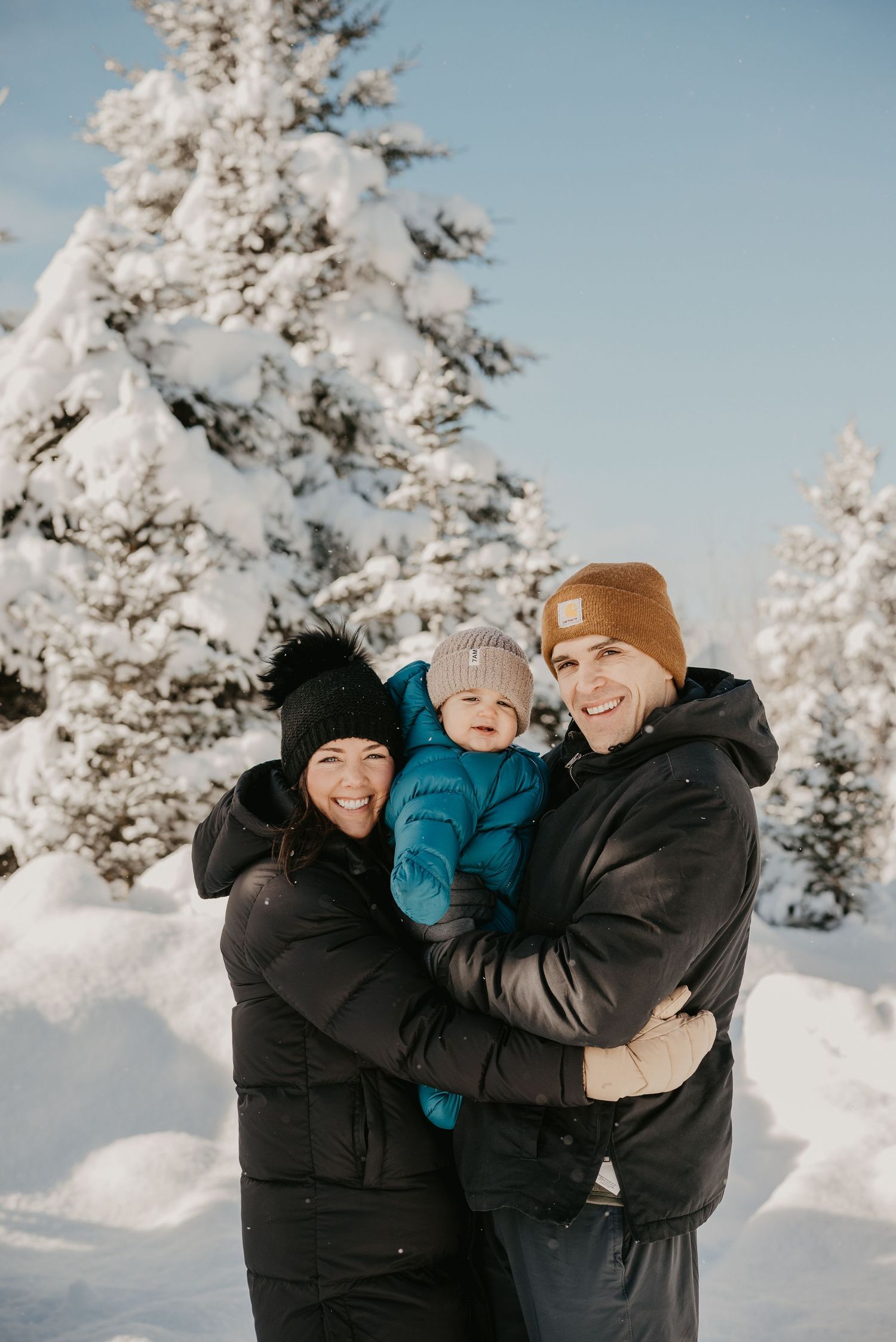 Happy family with baby in blue snowsuit poses among snow-covered pine trees in winter.