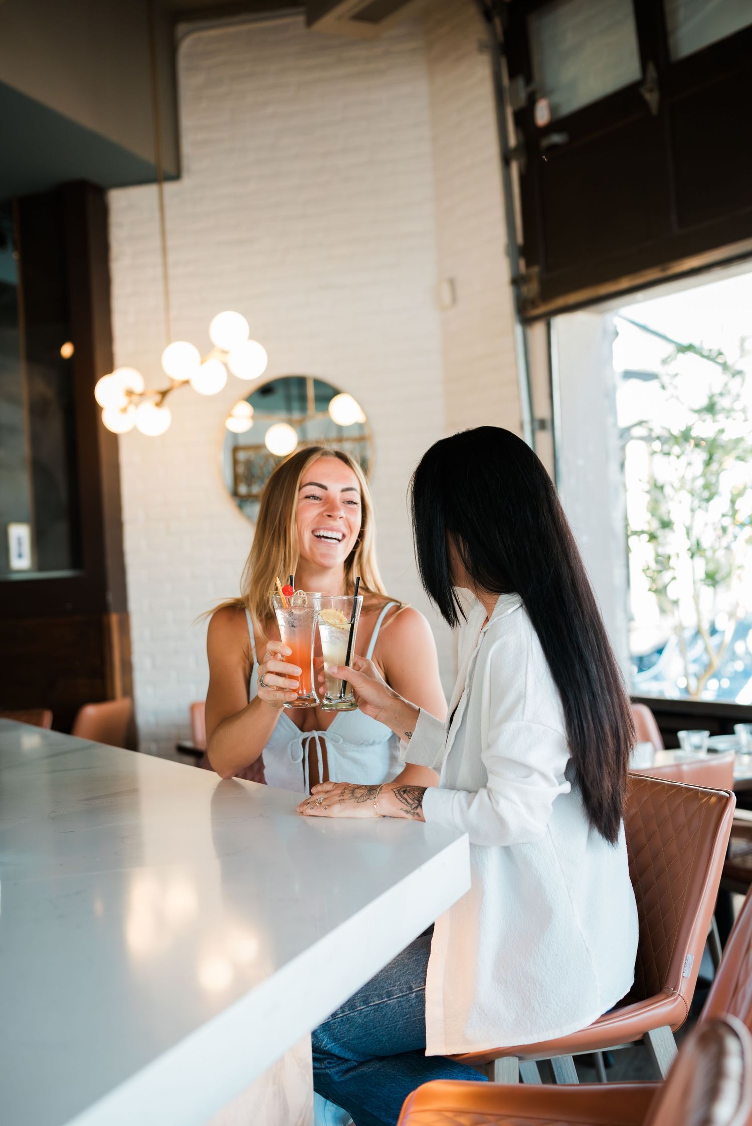 Two women laughing and clinking cocktail glasses together at a bright modern bar.