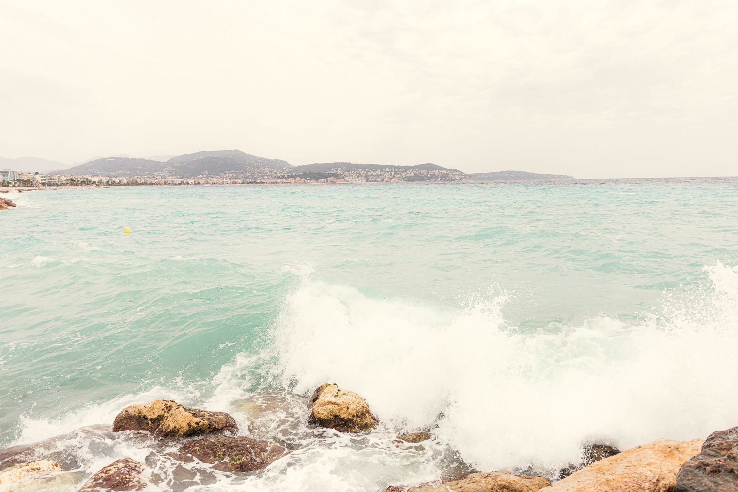 Waves crashing on rocky shoreline with turquoise Mediterranean Sea and distant hills under overcast sky.