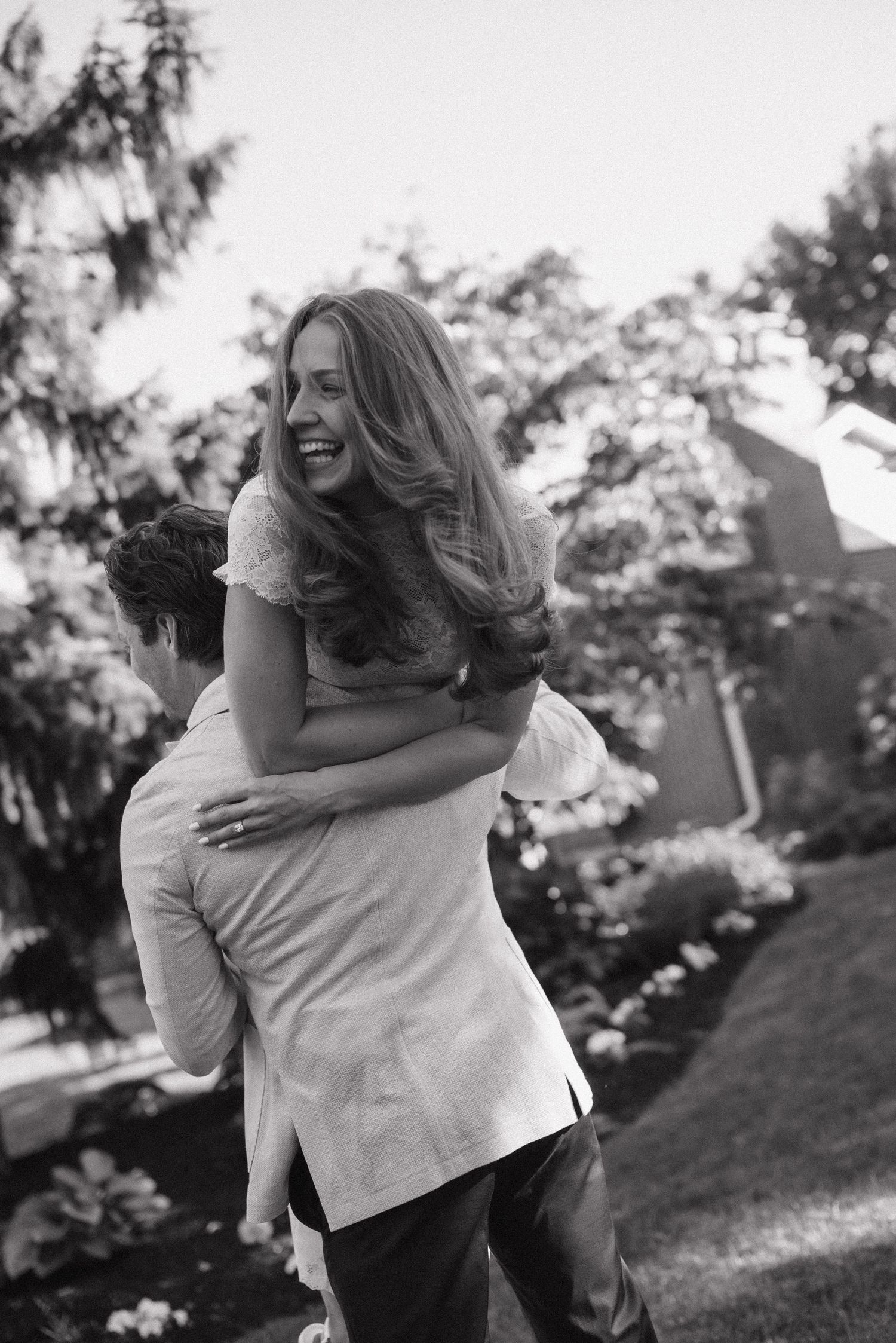 A joyful woman with long hair laughs while getting a piggyback ride outdoors in a garden setting, black and white photo.